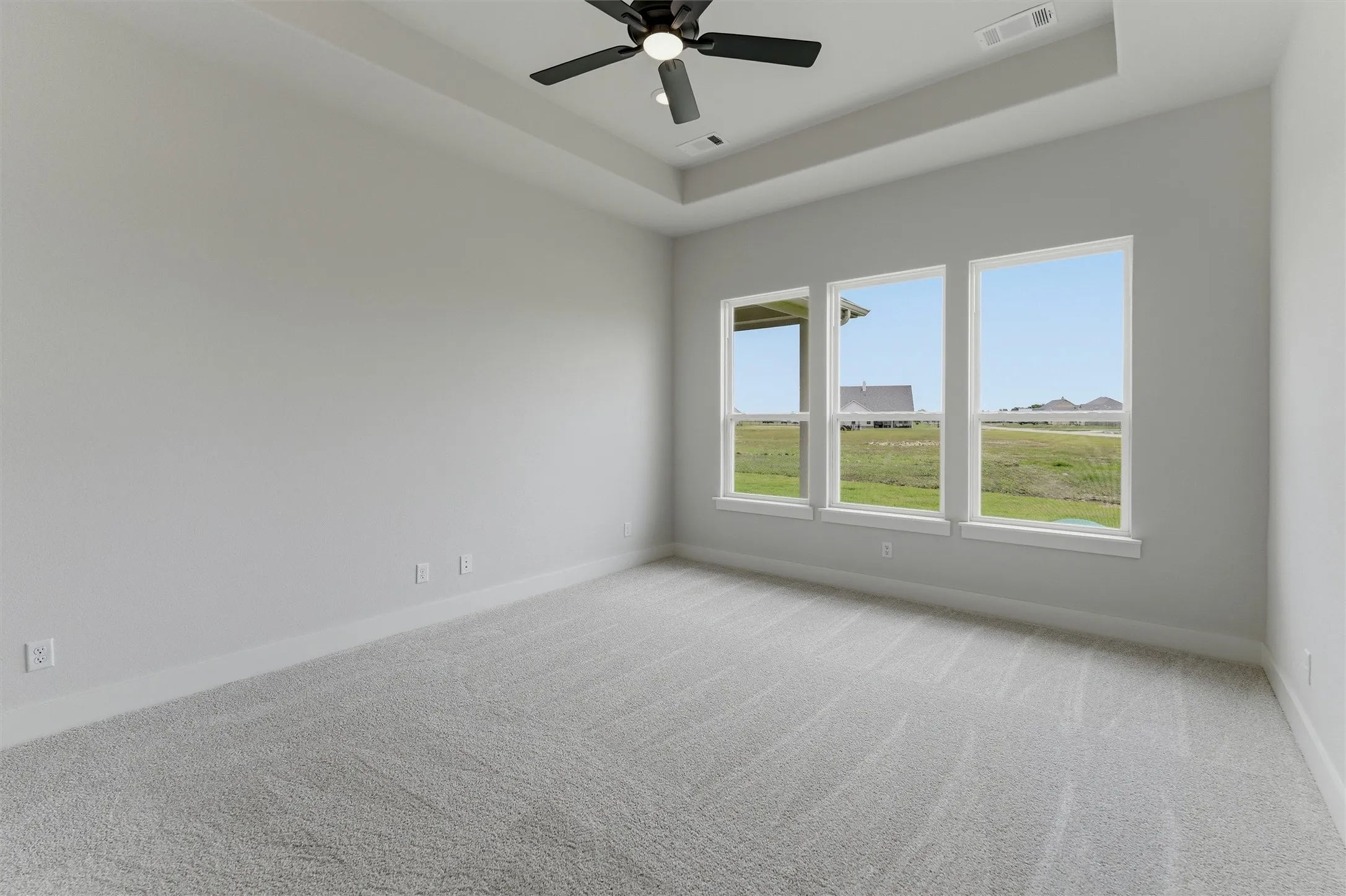 Expansive room featuring a tray ceiling with a black ceiling fan, light gray carpeting, and three large windows overlooking open green space