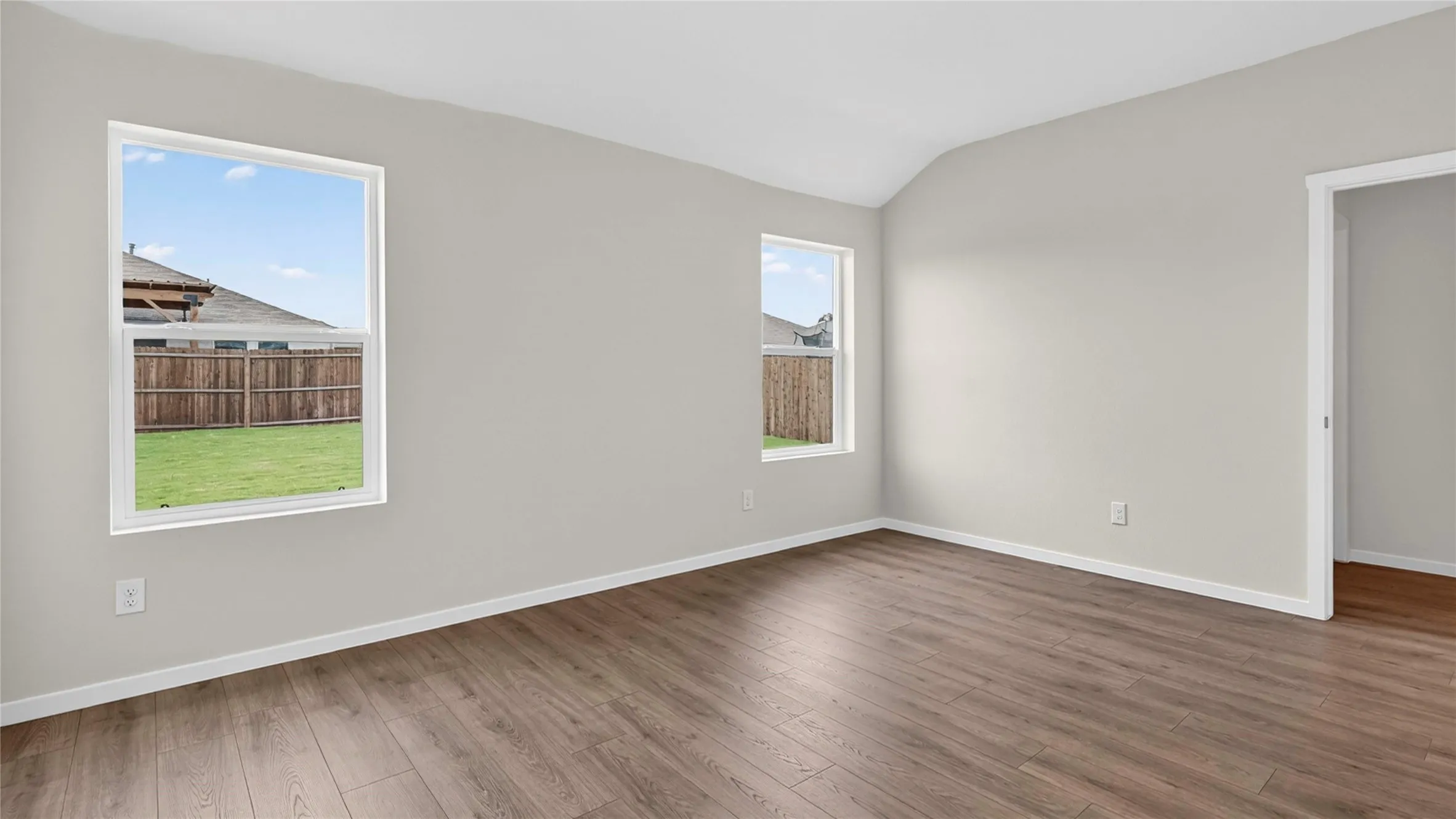 Spacious room featuring vaulted ceilings, two windows, wood-finish flooring, light gray wall paint, and white baseboards
