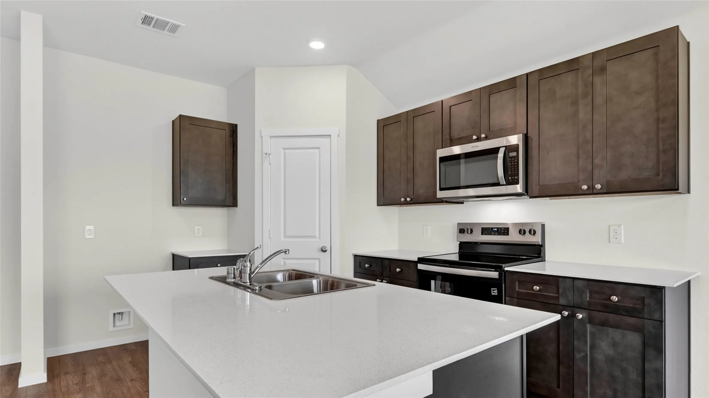 Kitchen featuring a center island with double basin sink, dark wood cabinetry, white countertops, stainless steel microwave, and wood-finish flooring