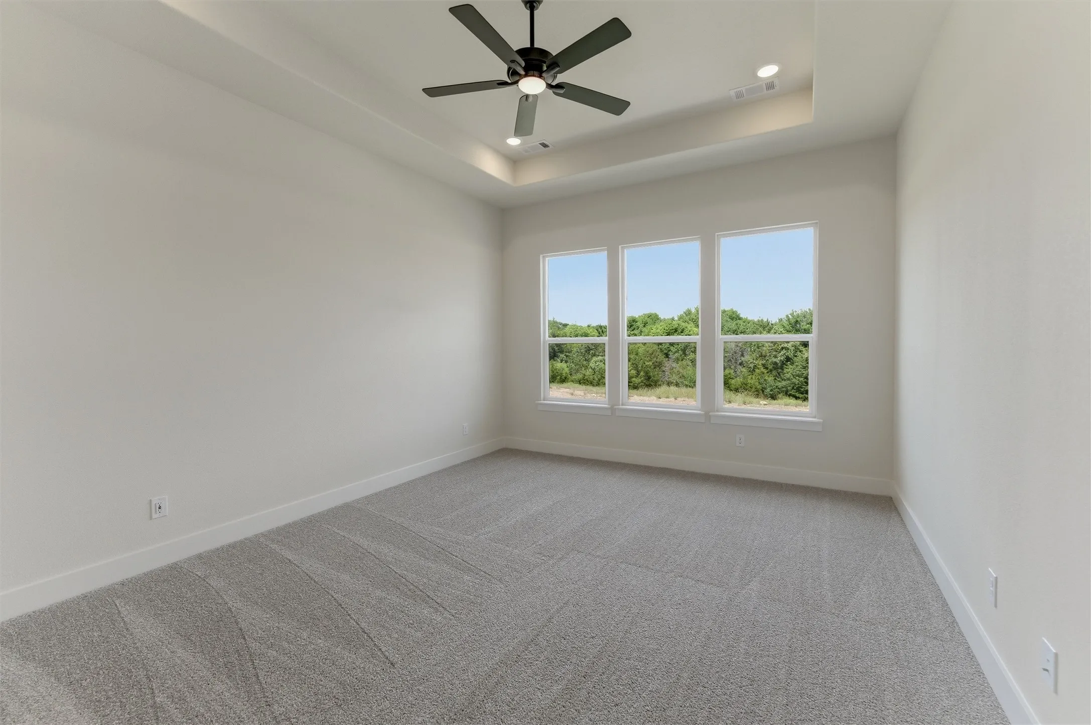 Room featuring a tray ceiling with recessed lighting and a ceiling fan, three large windows, light gray patterned carpeting, and white baseboards