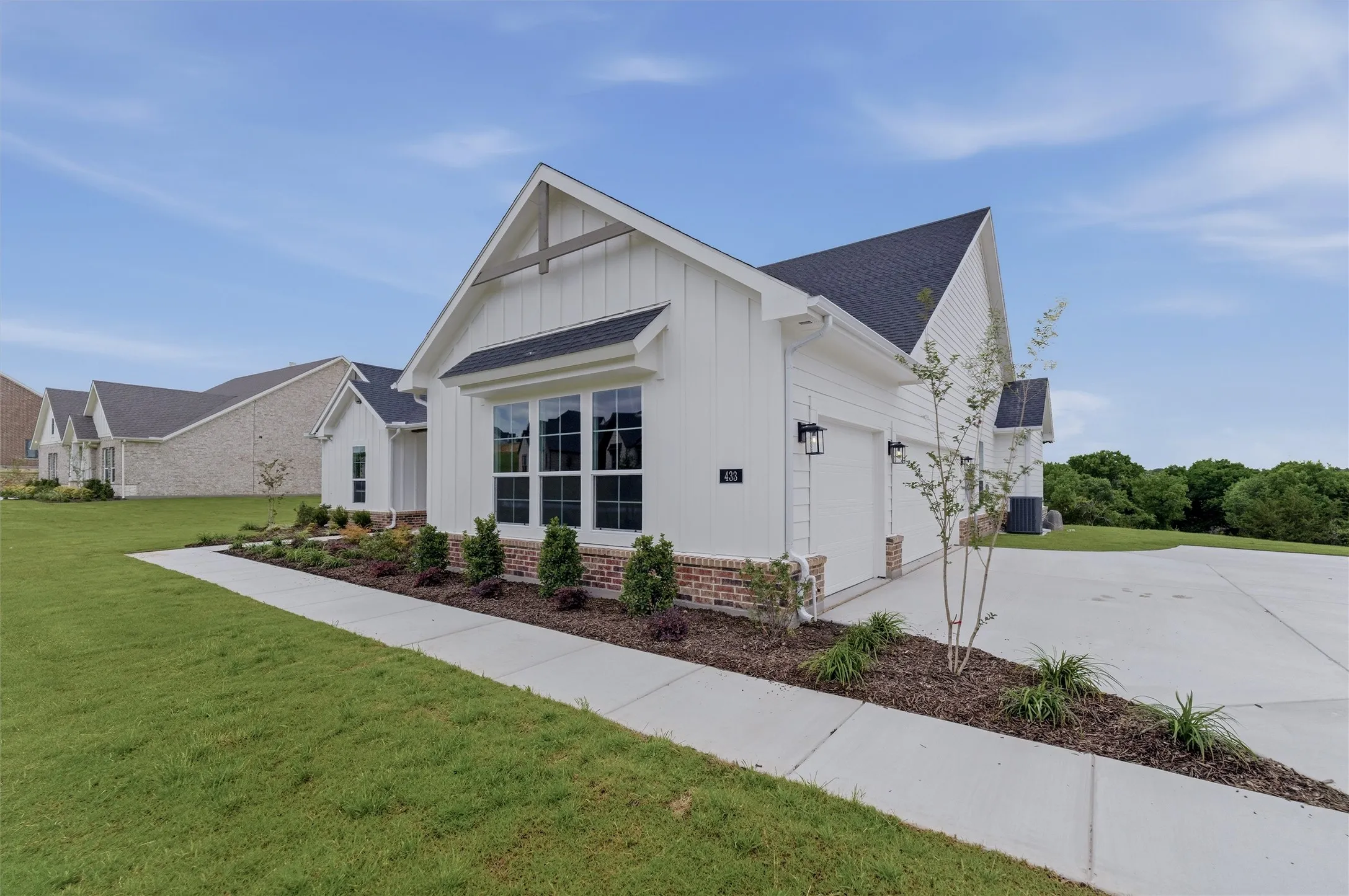 Modern farmhouse exterior featuring white board and batten siding, dark shingle roofing, a brick foundation, a concrete driveway, and an established lawn with landscaping