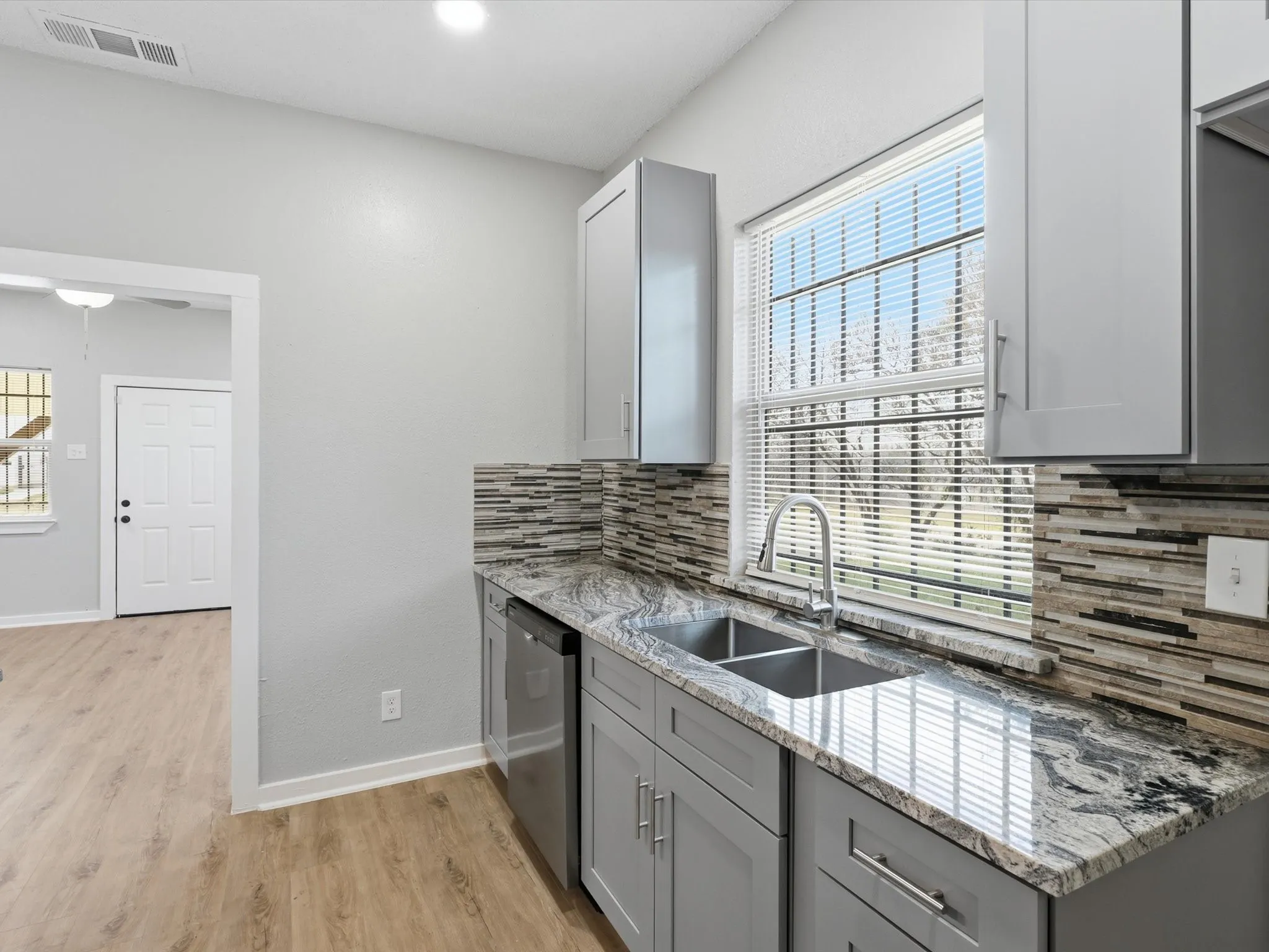 Kitchen featuring gray cabinetry, light stone counters, tasteful backsplash, stainless steel dishwasher, and light wood finished floors