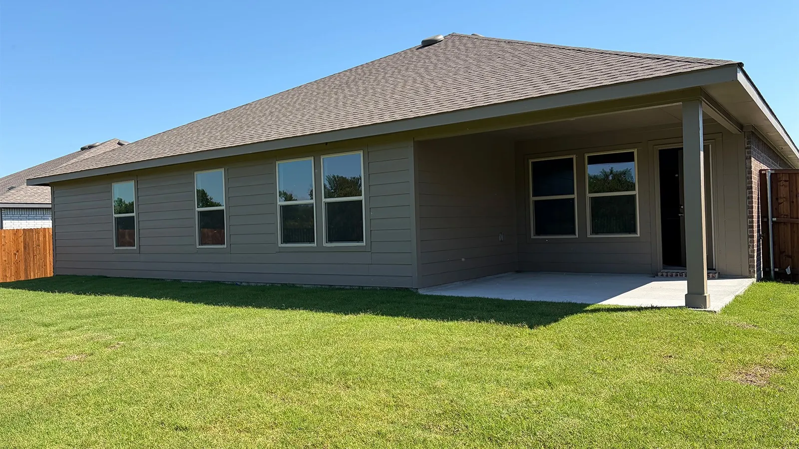 Rear exterior featuring a covered concrete patio, grey horizontal siding, numerous windows, a brown shingle roof, and a wood fence
