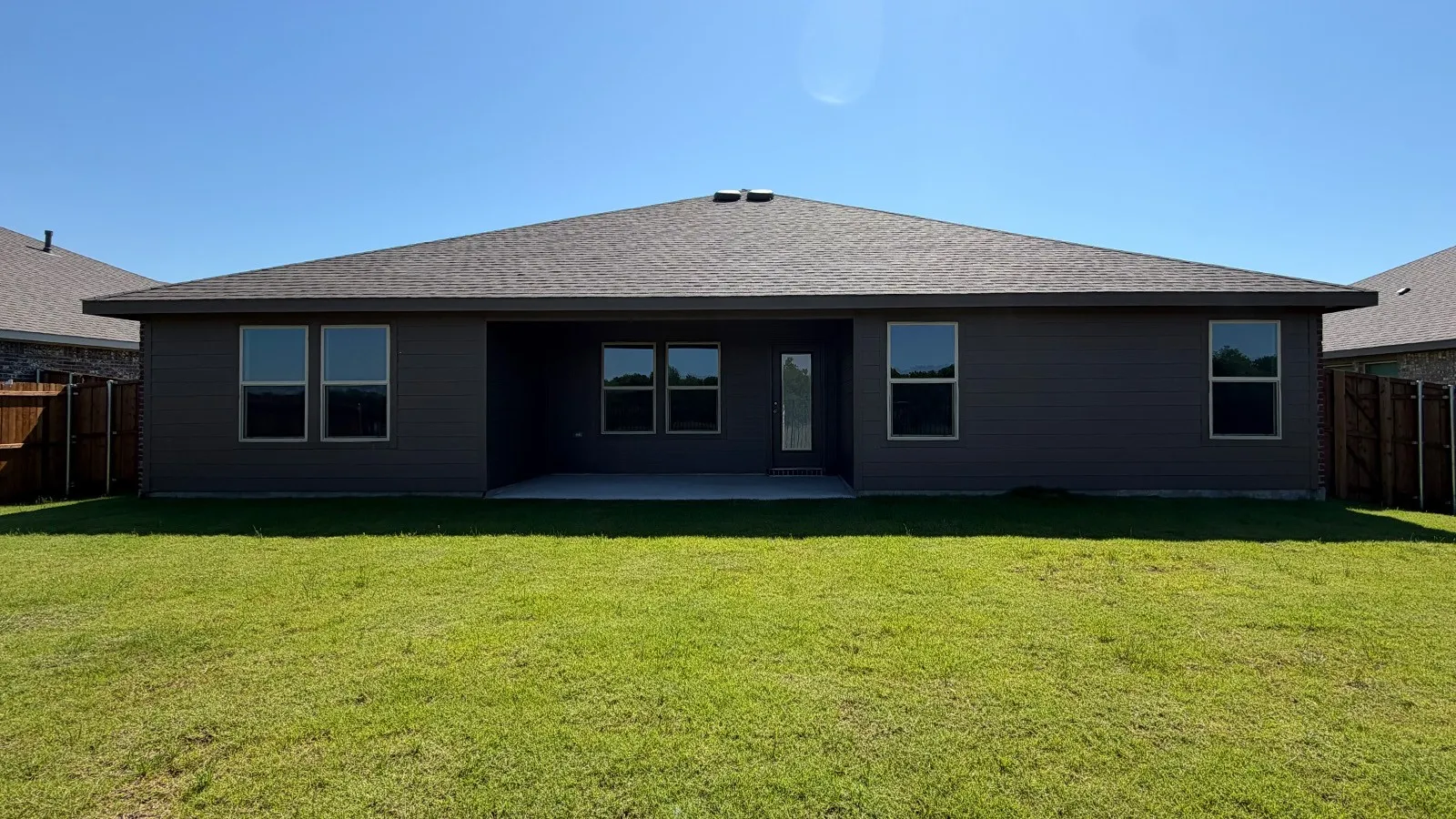 Expansive lawn featuring a concrete patio, dark gray siding, multiple windows, and a shingled roof