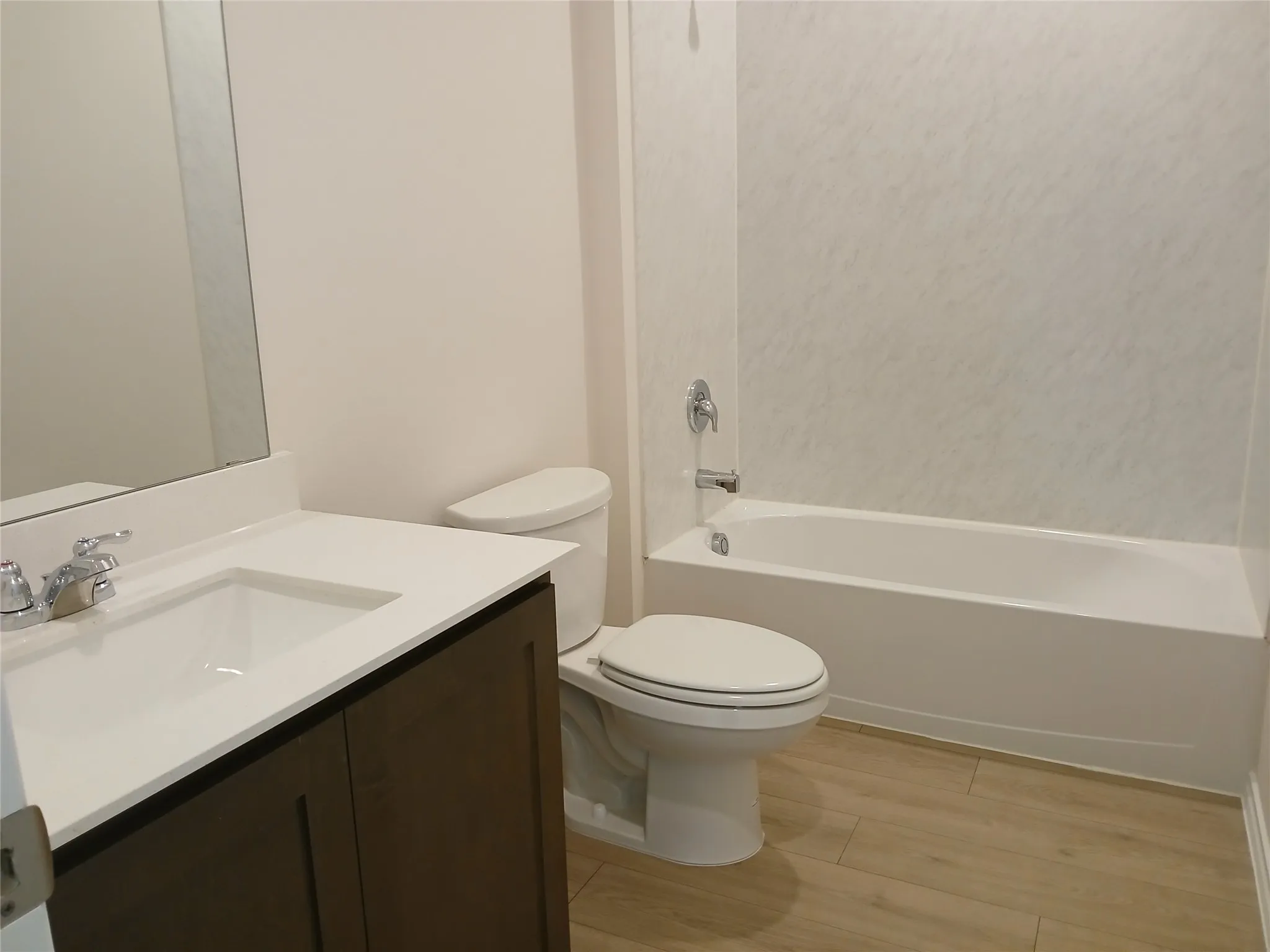 Bathroom featuring a dark wood-finish vanity with a white countertop and integrated sink, a full-width mirror, a white toilet, a white bathtub with a shower surround, and wood-finish flooring