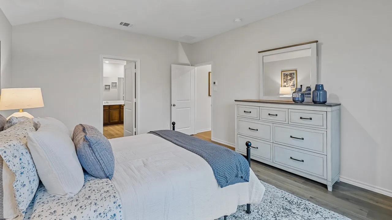 Spacious bedroom featuring light neutral walls and wood-finish flooring, with an open doorway revealing a bathroom with a vanity and wood-finish cabinetry