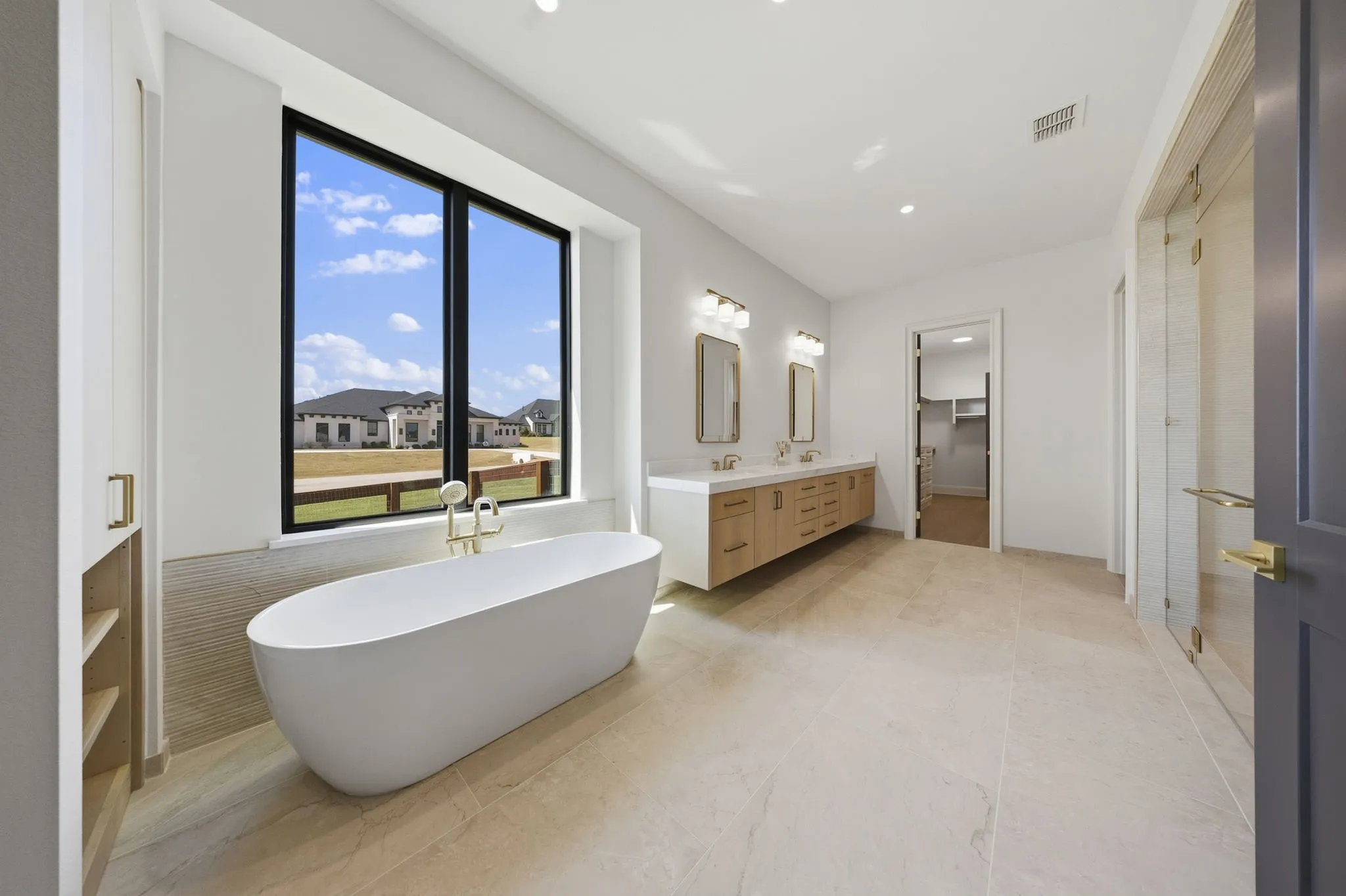 Freestanding soaking tub with gold-finish fixtures, dual vanity with wood-finish cabinetry, and a large window with black trim