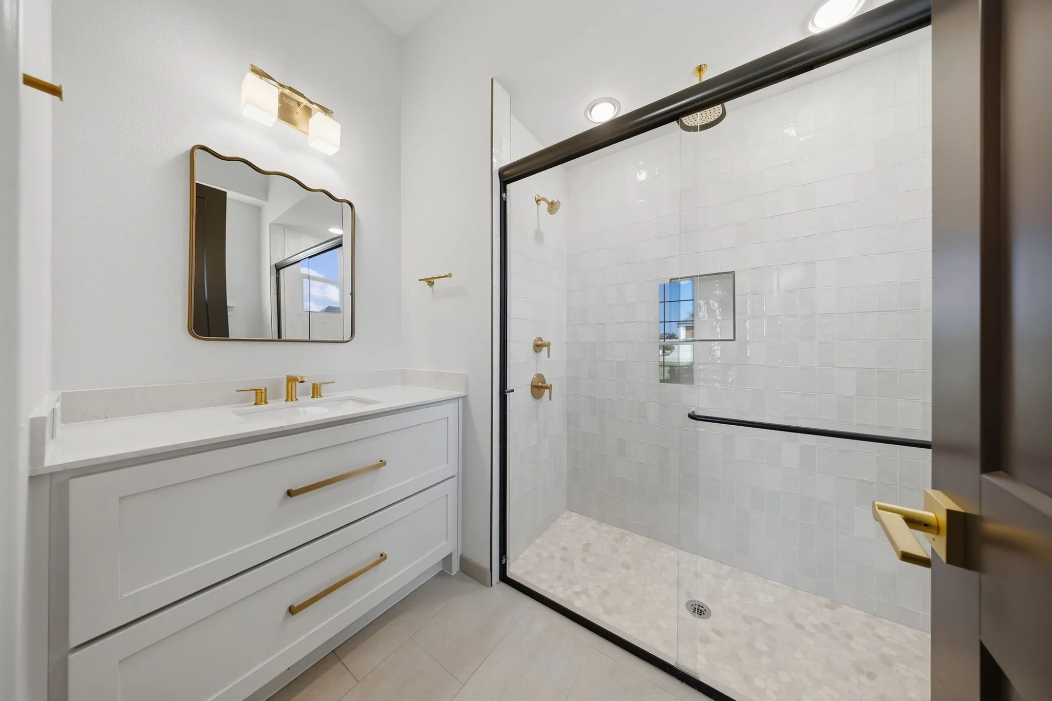 White shaker vanity with quartz countertop, gold-finish hardware, and a frameless glass shower enclosure featuring a tiled interior