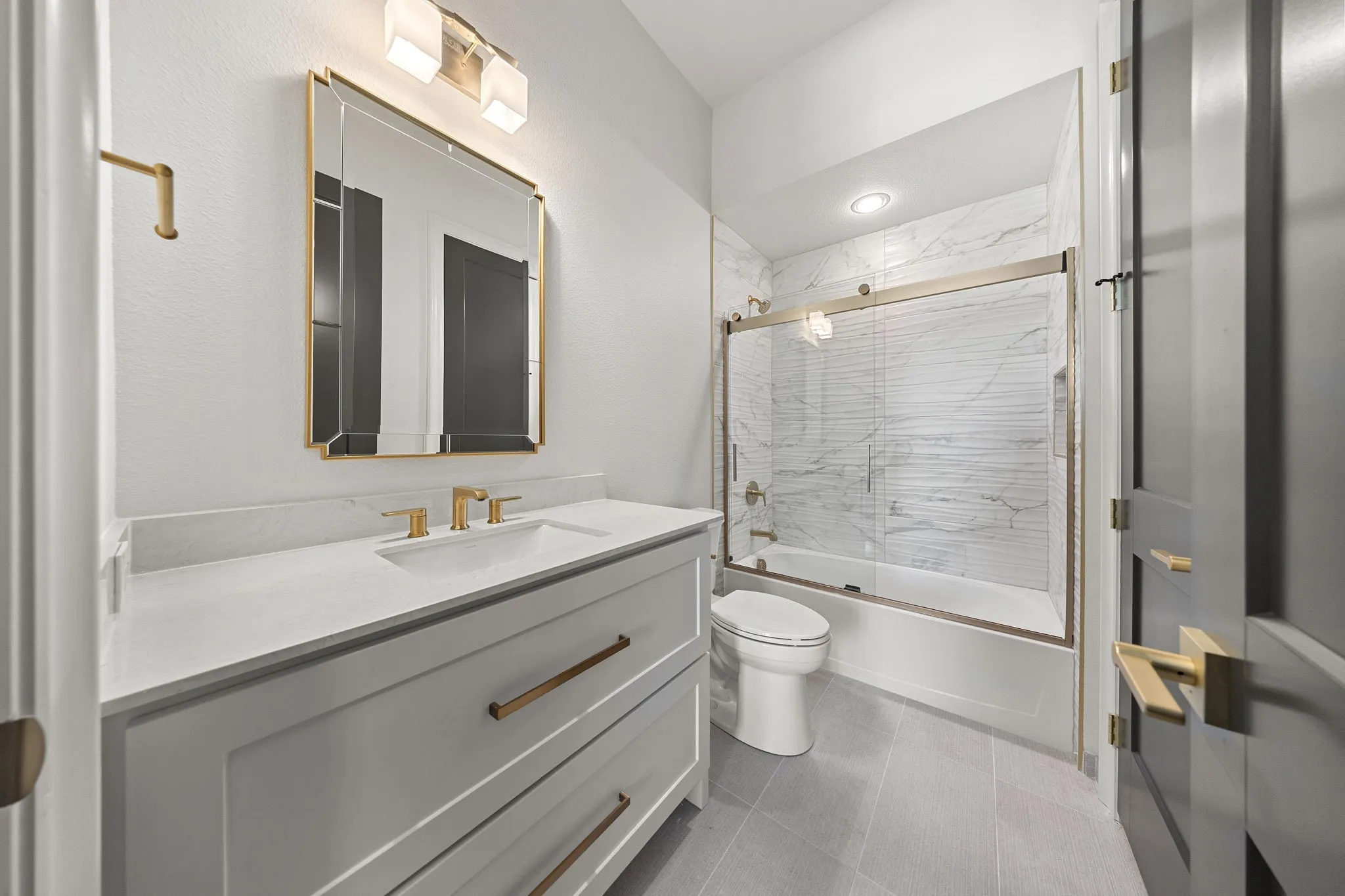 White shaker vanity with white countertop, gold-finish hardware, gold-finish framed mirror, and a white tiled shower with sliding glass doors