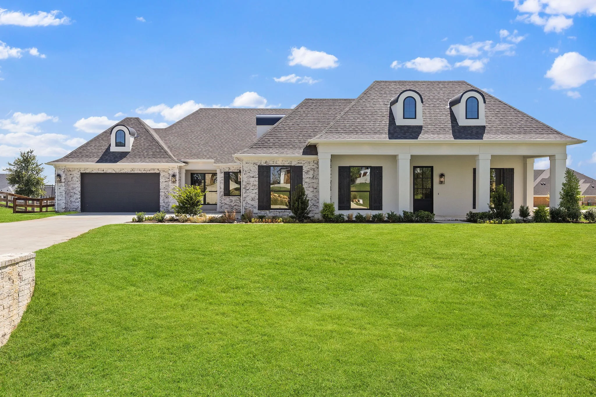 Exterior featuring a combination of brick and stucco finishes, covered front porch with substantial columns, a dark shingle roof, arched dormer windows, and an integrated garage with a dark panel door