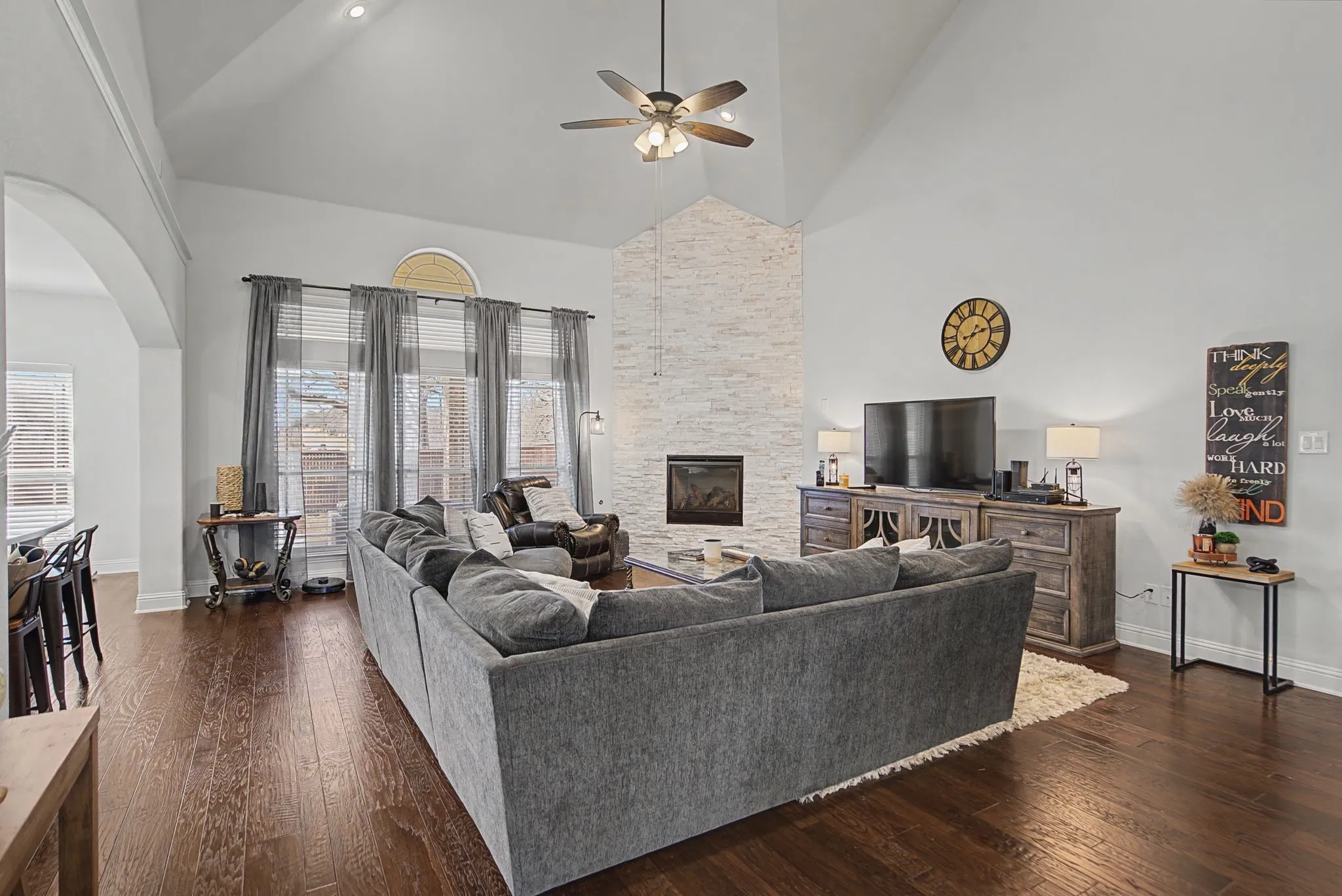 Living area with dark wood finished floors, a floor to ceiling stone fireplace, a ceiling fan, arched walkways, and vaulted ceiling