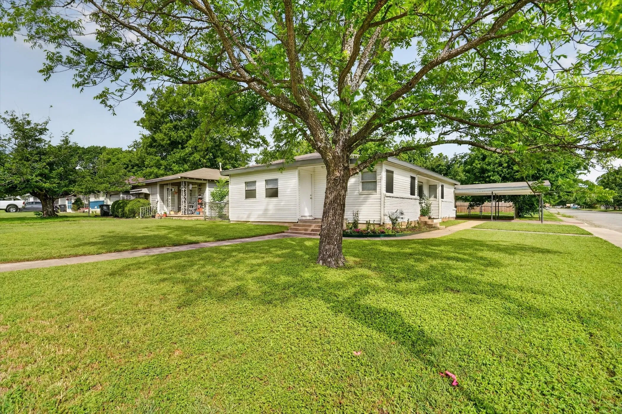 White siding residence with a covered carport, mature trees, a front lawn, and a sidewalk