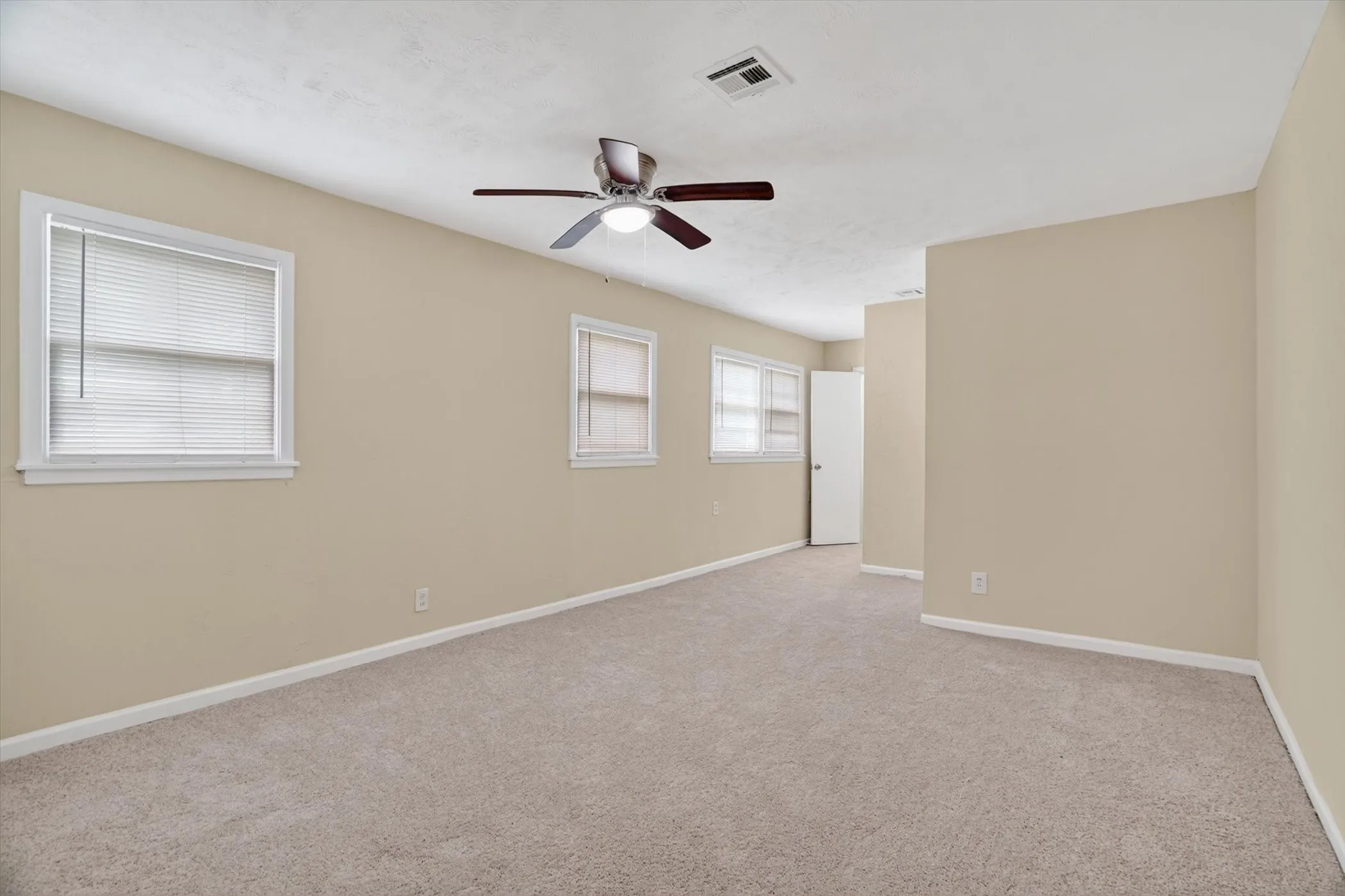 Spacious room featuring neutral carpeting, light beige wall paint, two white-framed windows with blinds, a ceiling fan with integrated lighting, and white baseboards