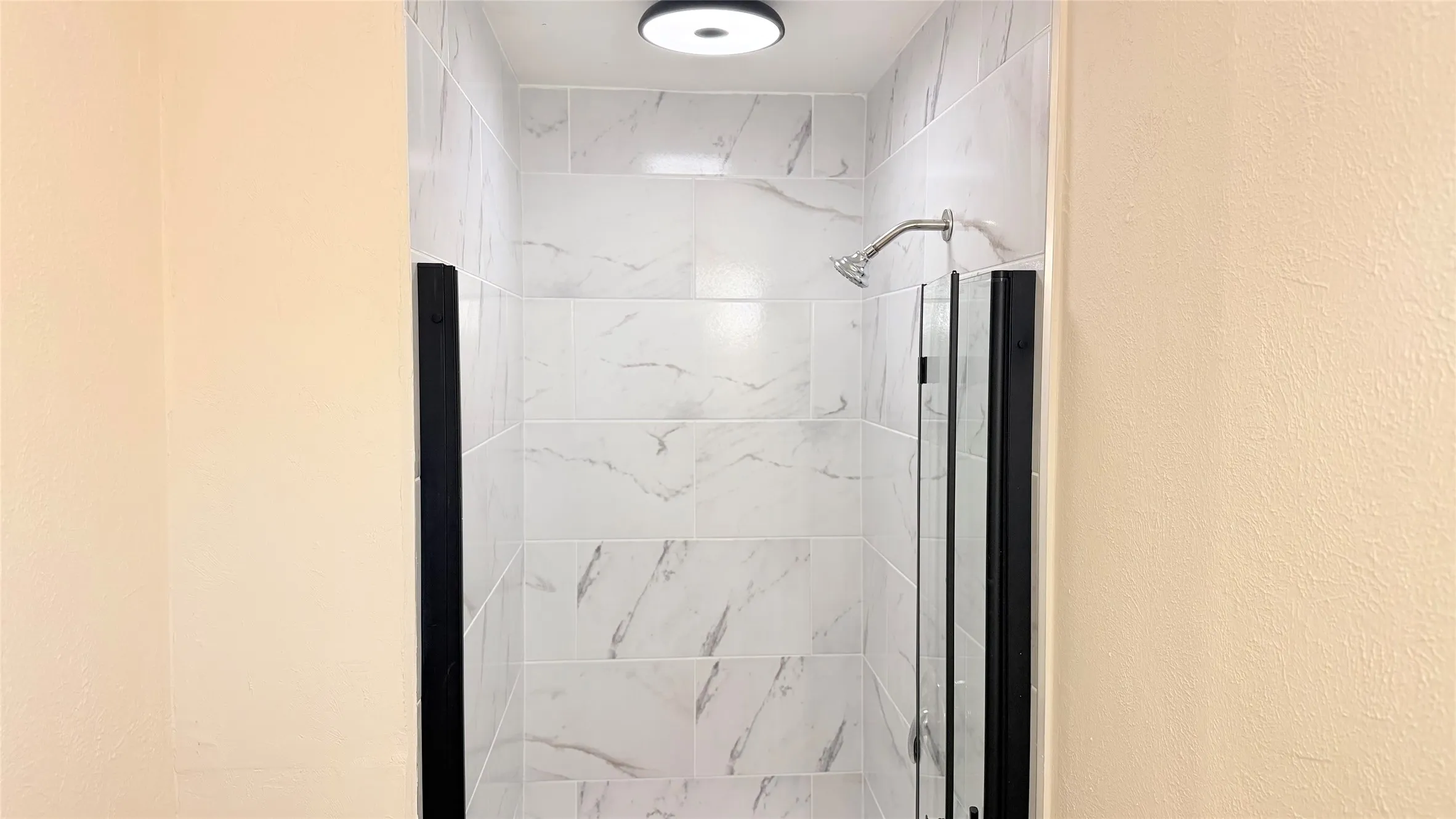 Shower enclosure featuring white marble-look subway tile, a chrome shower head, clear glass door panels with black framing, and a ceiling-mounted flush light fixture