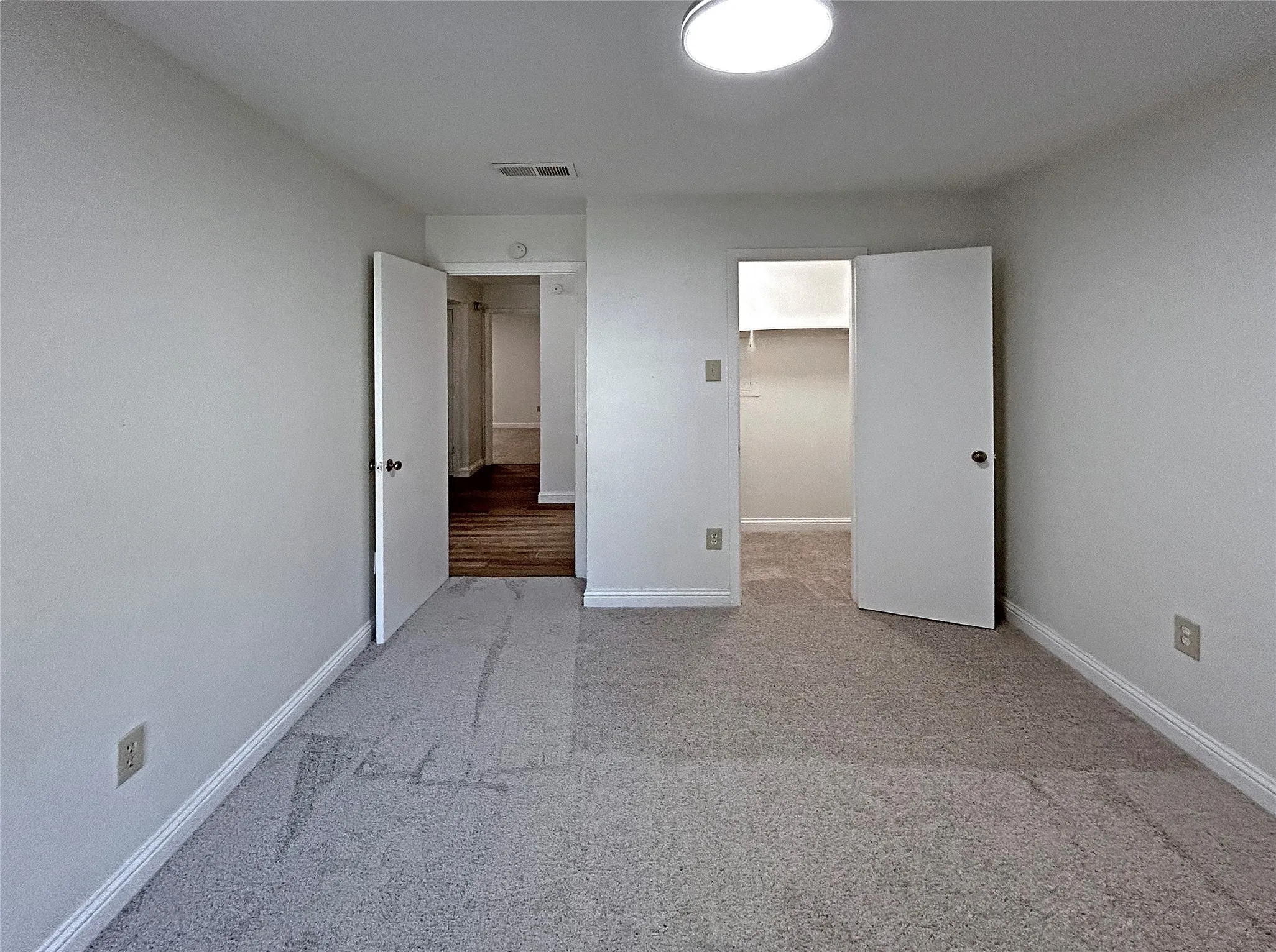 The secondary bedroom has light grey wall paint with white trim, new grey carpeting, ceiling-mounted light fixture, and white interior doors. It is located beside the second bathroom and has a front door view.