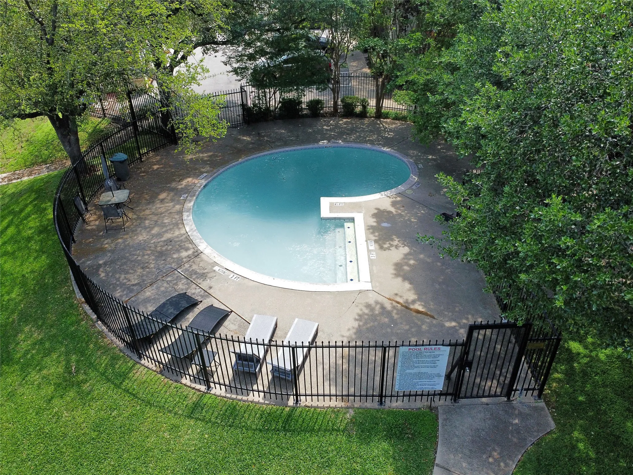 The freeform community swimming pool has integrated steps. It is surrounded by a concrete patio and black wrought iron fencing. Lounge chairs and a tables are available for residents.