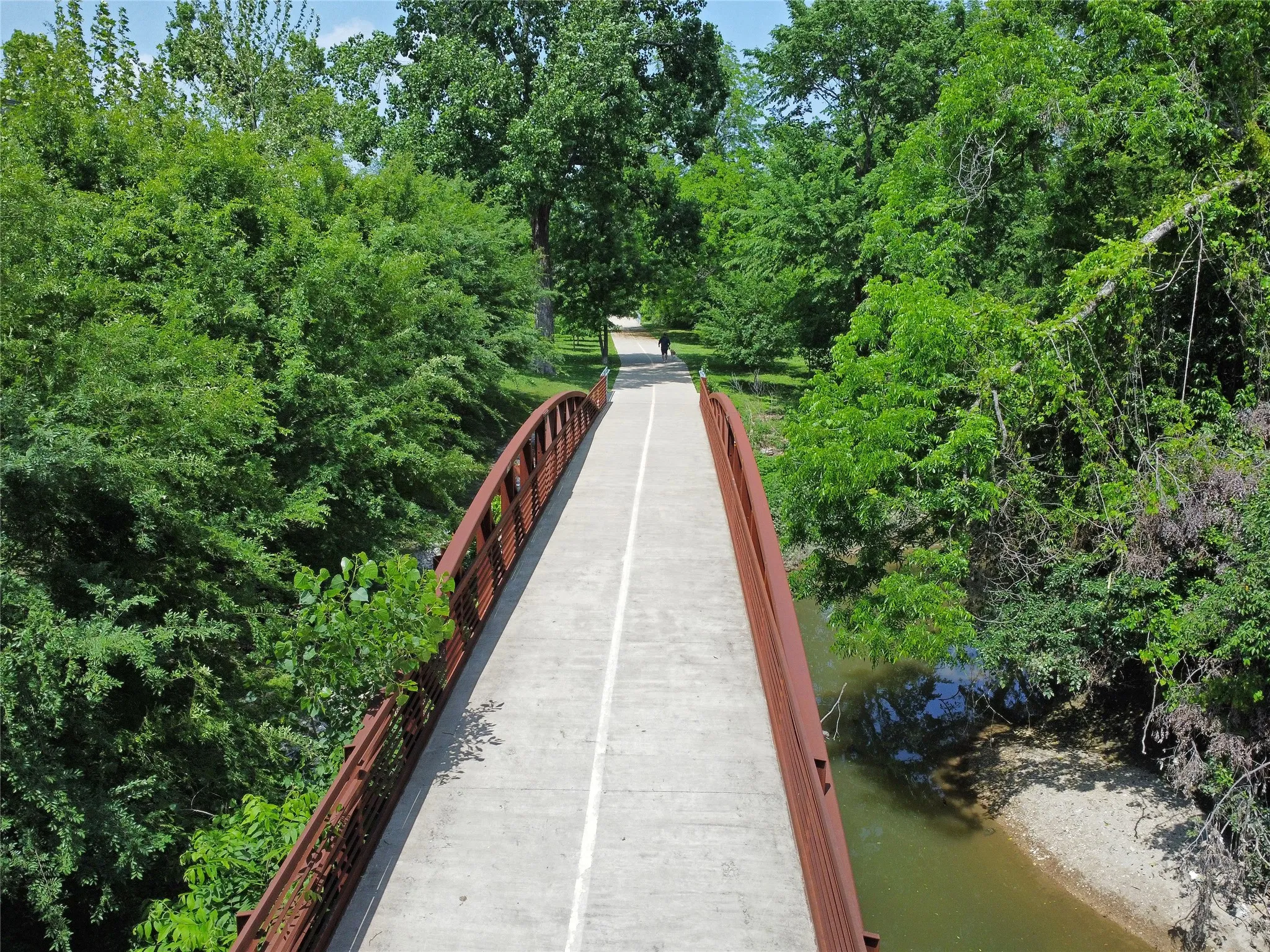 A bridge on Lake Highlands Trail is near the community pool at the rear of the property. The concrete hike & bike bridge has brown metal railings, surrounded by lush green foliage, extending over a bend in the natural waterway.