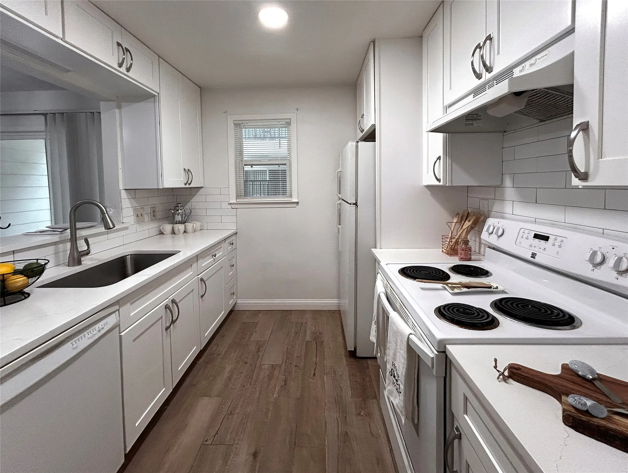 The kitchen has a window, breakfast bar, stainless steel sink, white appliances, white cabinetry, designer cabinets and subway tile backsplash.