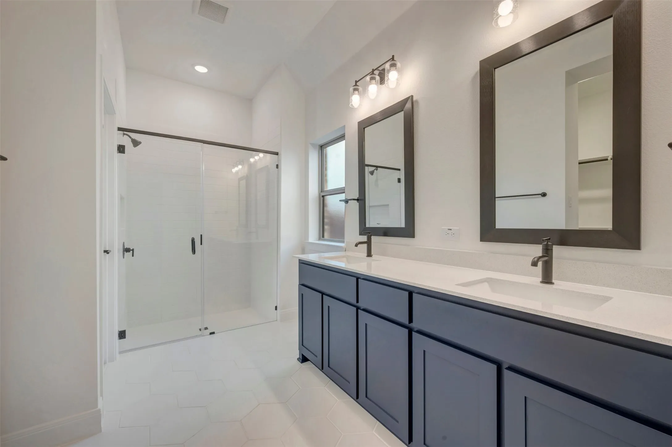 Spacious bathroom featuring a dual vanity with white countertops and dark cabinetry, individual framed mirrors, and a glass-enclosed shower with dark hardware