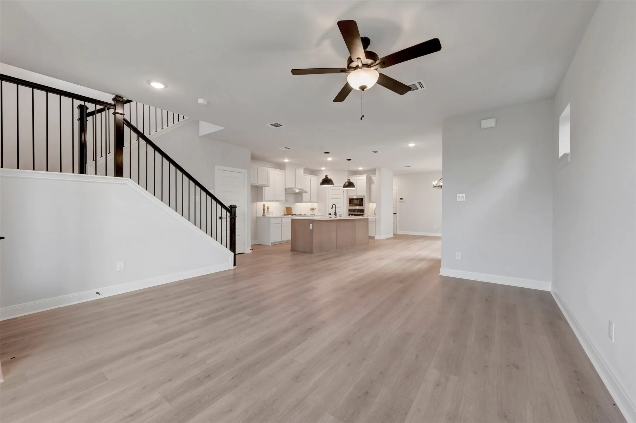 Open-concept interior featuring light wood-finish flooring, a contemporary ceiling fan, and a kitchen with white cabinetry, an island with pendant lighting, and stainless steel appliances