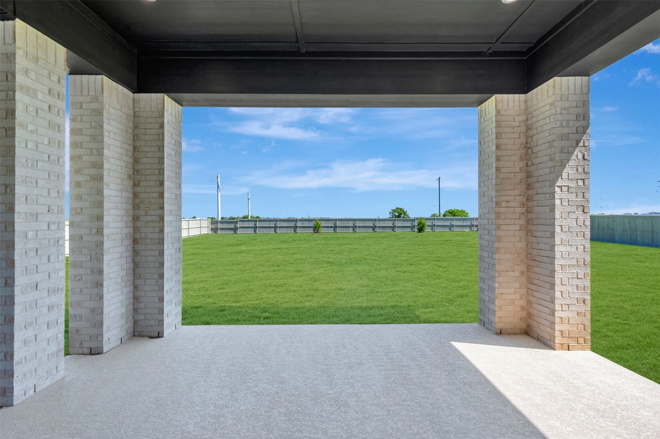 Covered patio with a light aggregate finish, supported by light-toned brick pillars, overlooking an expansive green lawn