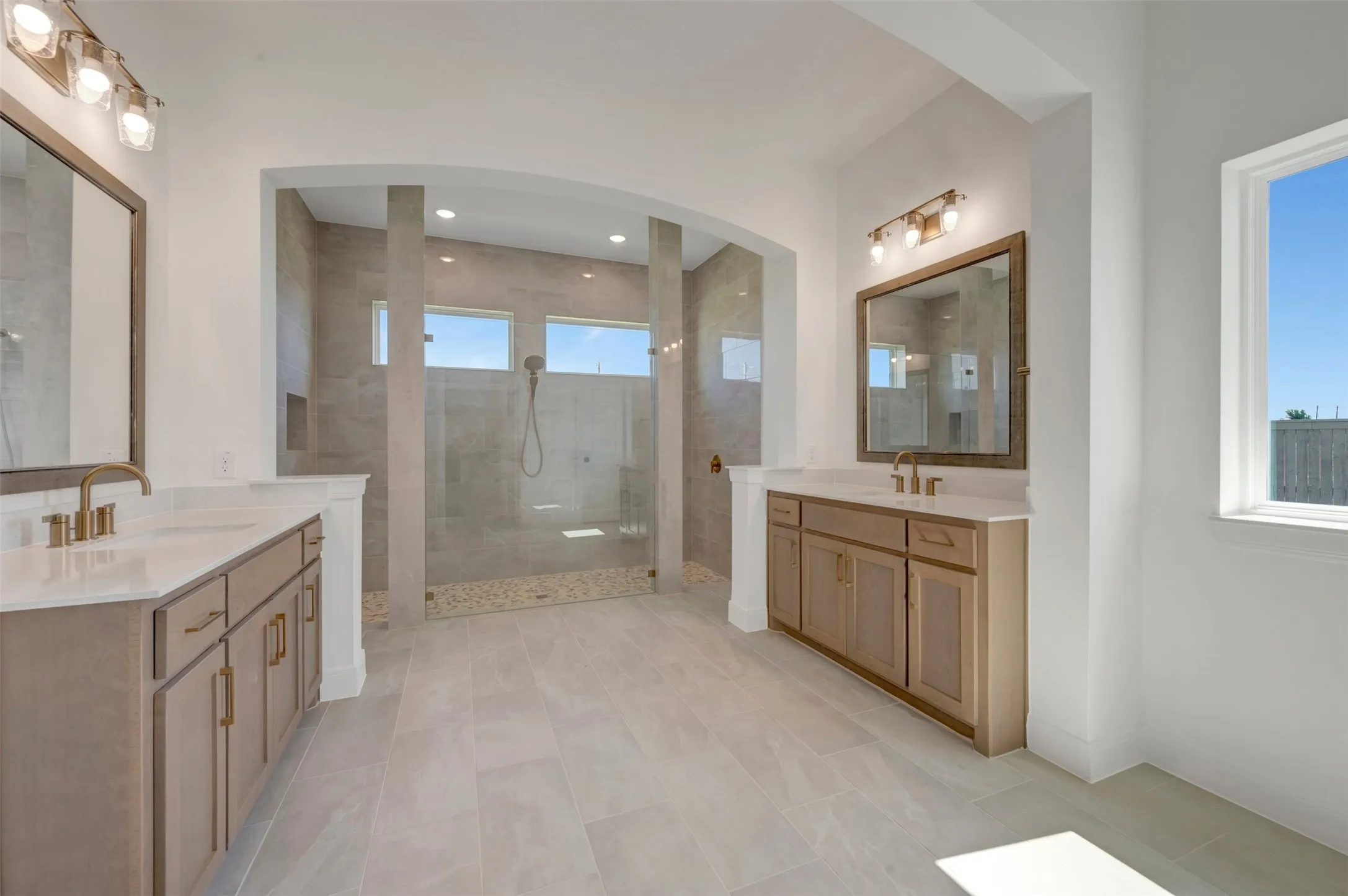 Spacious bathroom featuring a large walk-in shower with tiled walls and pebble-finish floor, dual vanities with light wood-finish cabinetry, white countertops, and bronze-finish fixtures