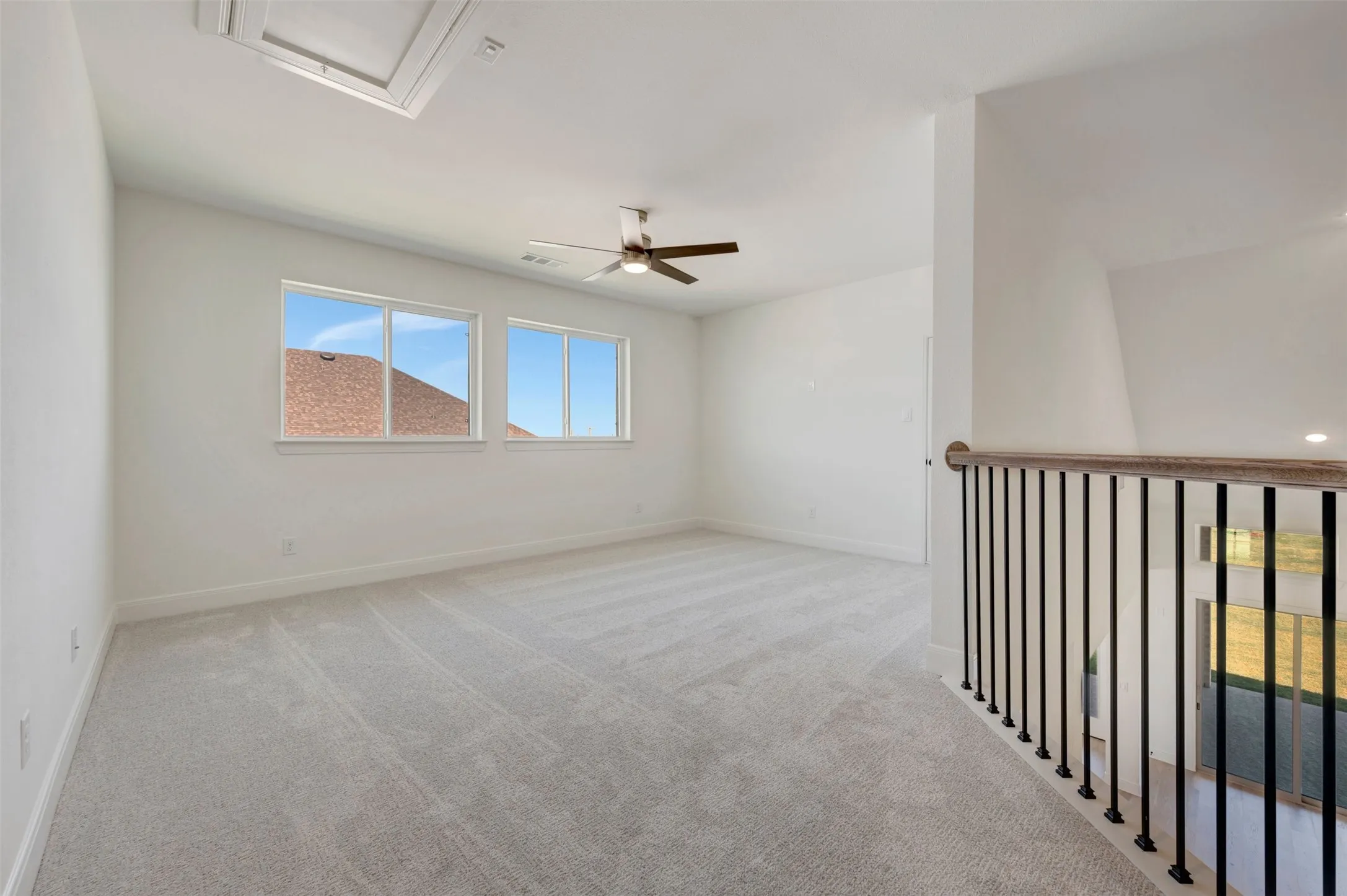 Loft space featuring light gray carpeting, crisp white walls, multiple windows, a ceiling fan, and a wood-finish handrail with black iron balusters
