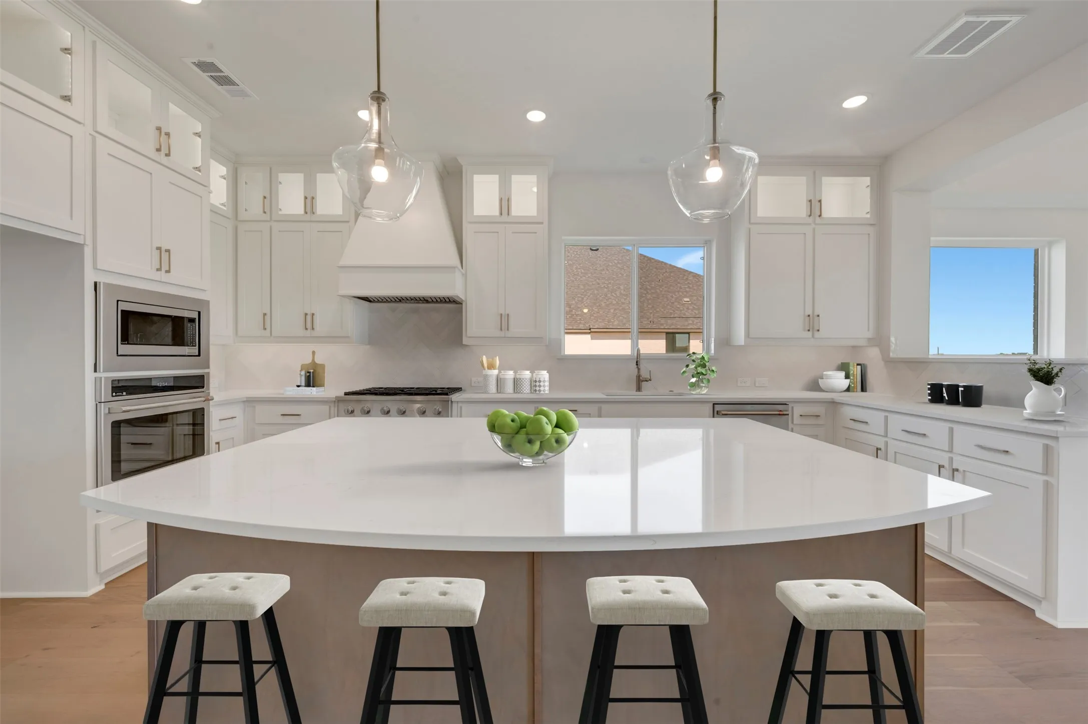 Spacious kitchen featuring a large central island with a light-toned countertop, white cabinetry with glass-front uppers, stainless steel built-in appliances, and light wood-finish flooring
