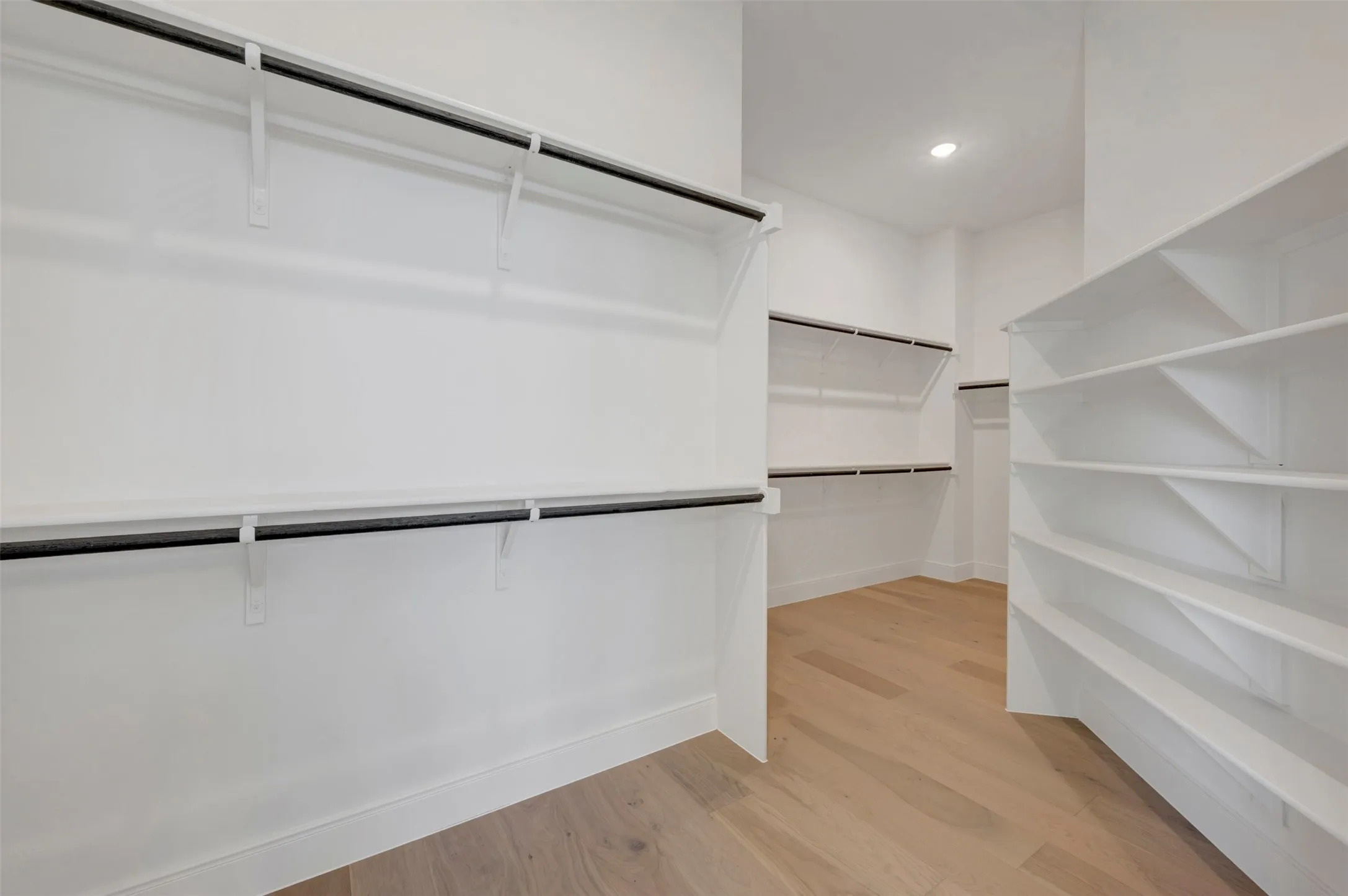 Walk-in closet featuring light wood-finish flooring, recessed lighting, white built-in shelving, and multiple hanging rods