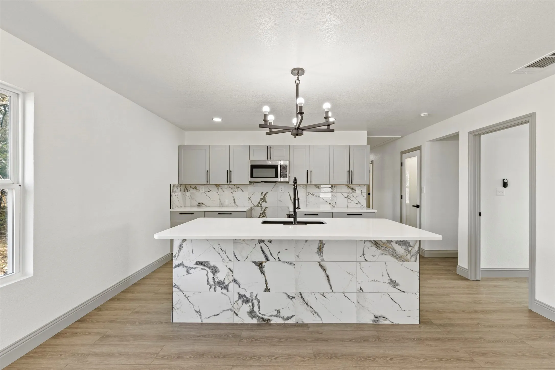 Kitchen featuring a center island with sink, light wood-type flooring, gray cabinetry, suspended lighting, and stainless steel microwave