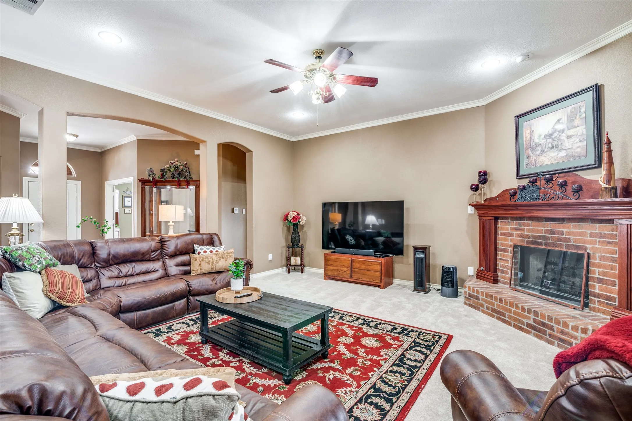 Carpeted living area featuring crown molding, ceiling fan, a brick fireplace, and arched walkways