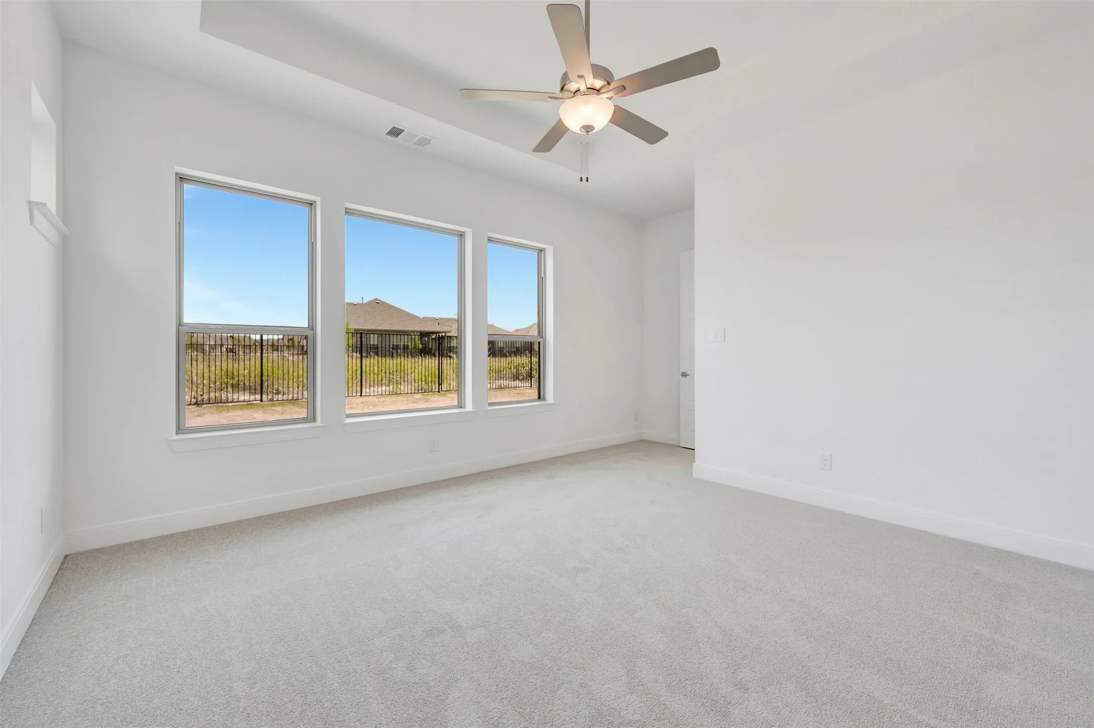 Bright interior space with light-toned carpeting, featuring a ceiling fan with integrated lighting, a tray ceiling detail, and three large windows providing ample natural illumination