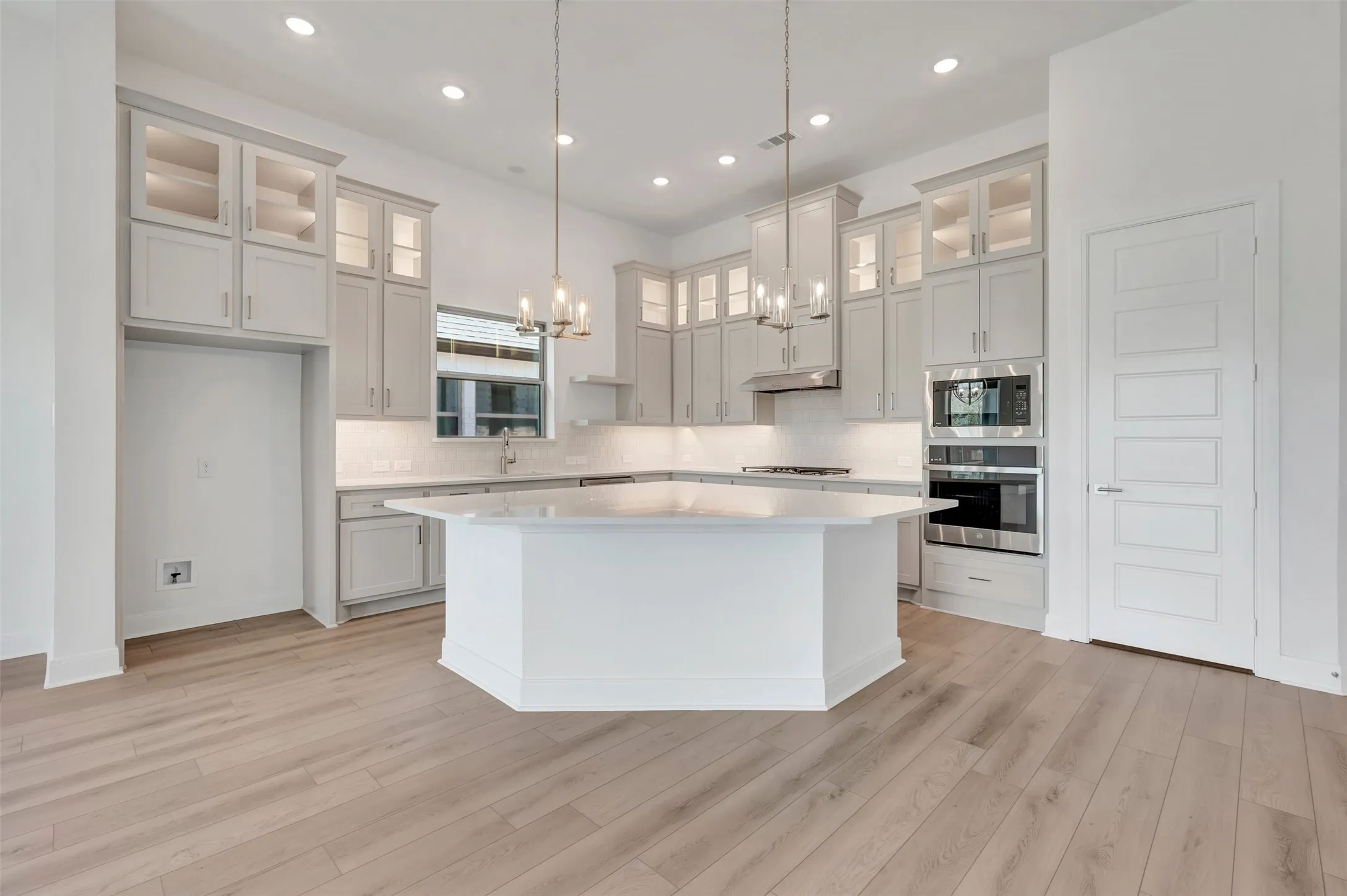 Open-concept kitchen featuring an oversized island, light-finish wood flooring, built-in stainless steel appliances, upper cabinetry with glass inserts, and recessed lighting