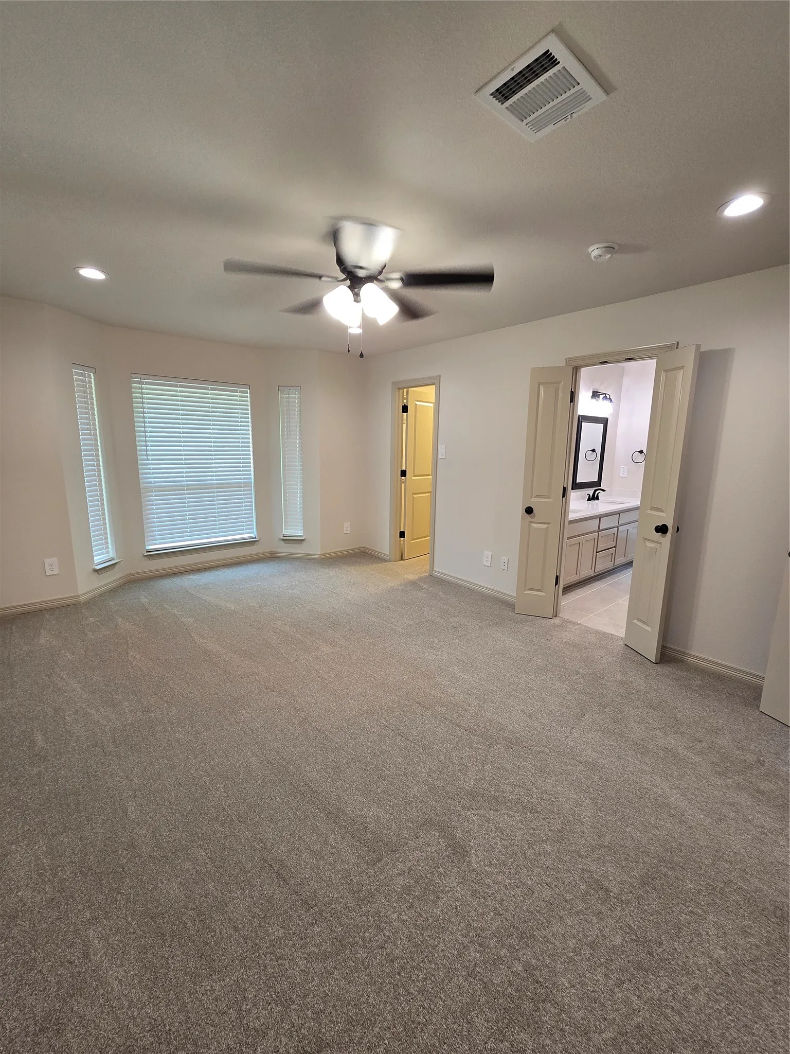 Primary bedroom featuring bay window, light colored carpet, recessed lighting, and a ceiling fan with doorways to closet and bathroom