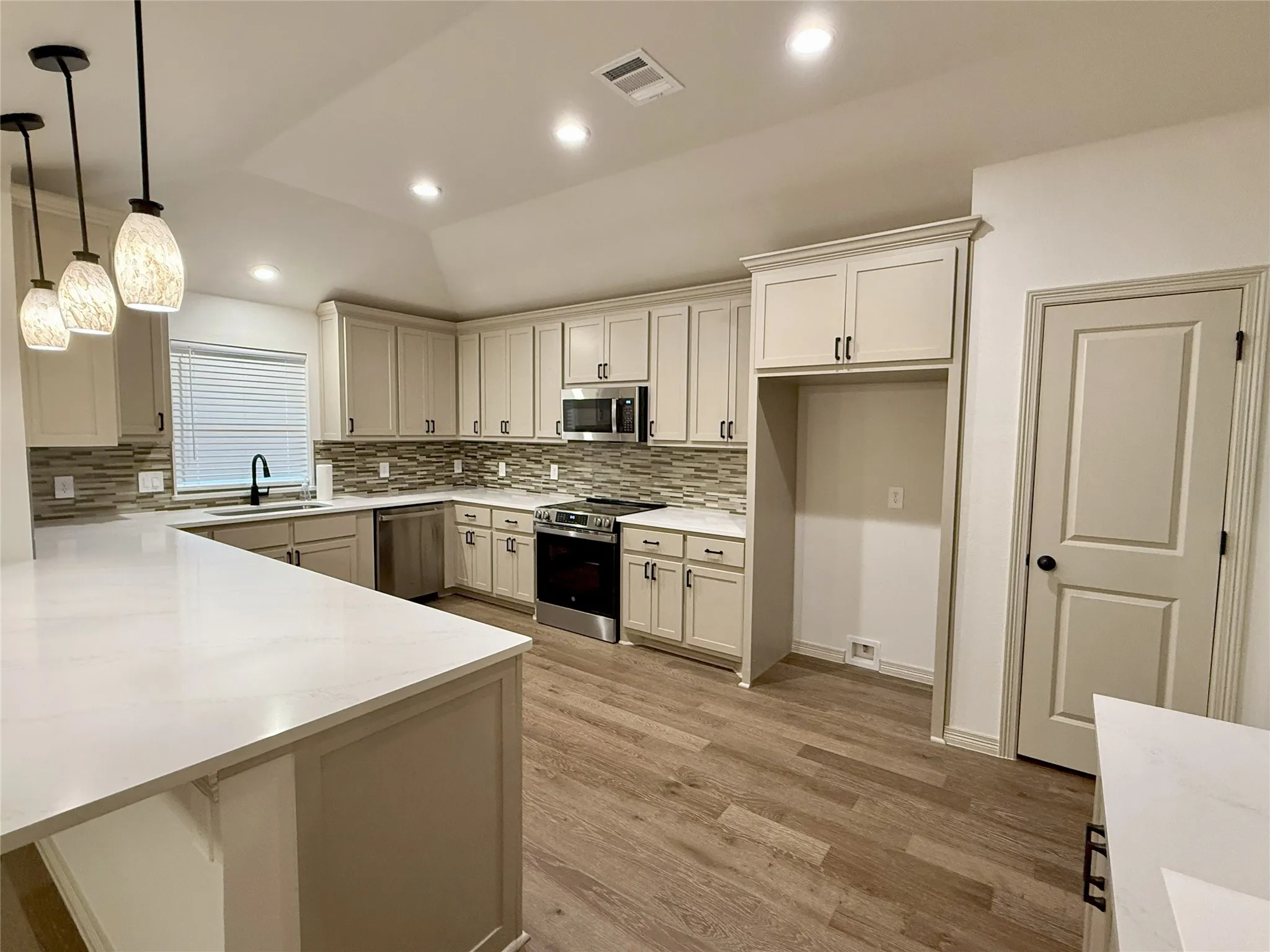 Kitchen featuring appliances with stainless steel finishes, light wood-style flooring, tasteful backsplash, a peninsula, custom cabinets and hanging light fixtures