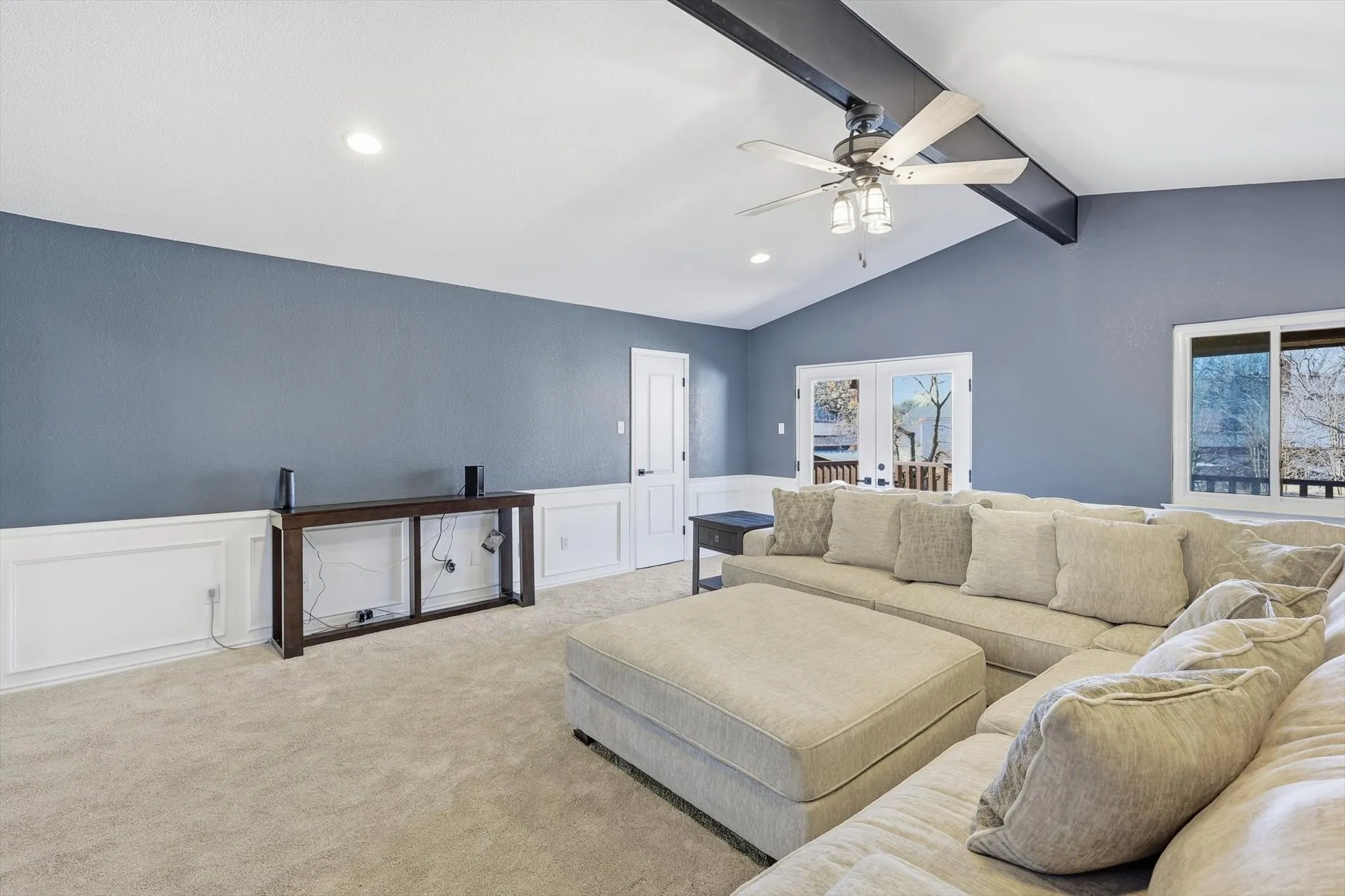 Living room featuring wainscoting, a decorative wall, ceiling fan, carpet flooring, and beamed ceiling