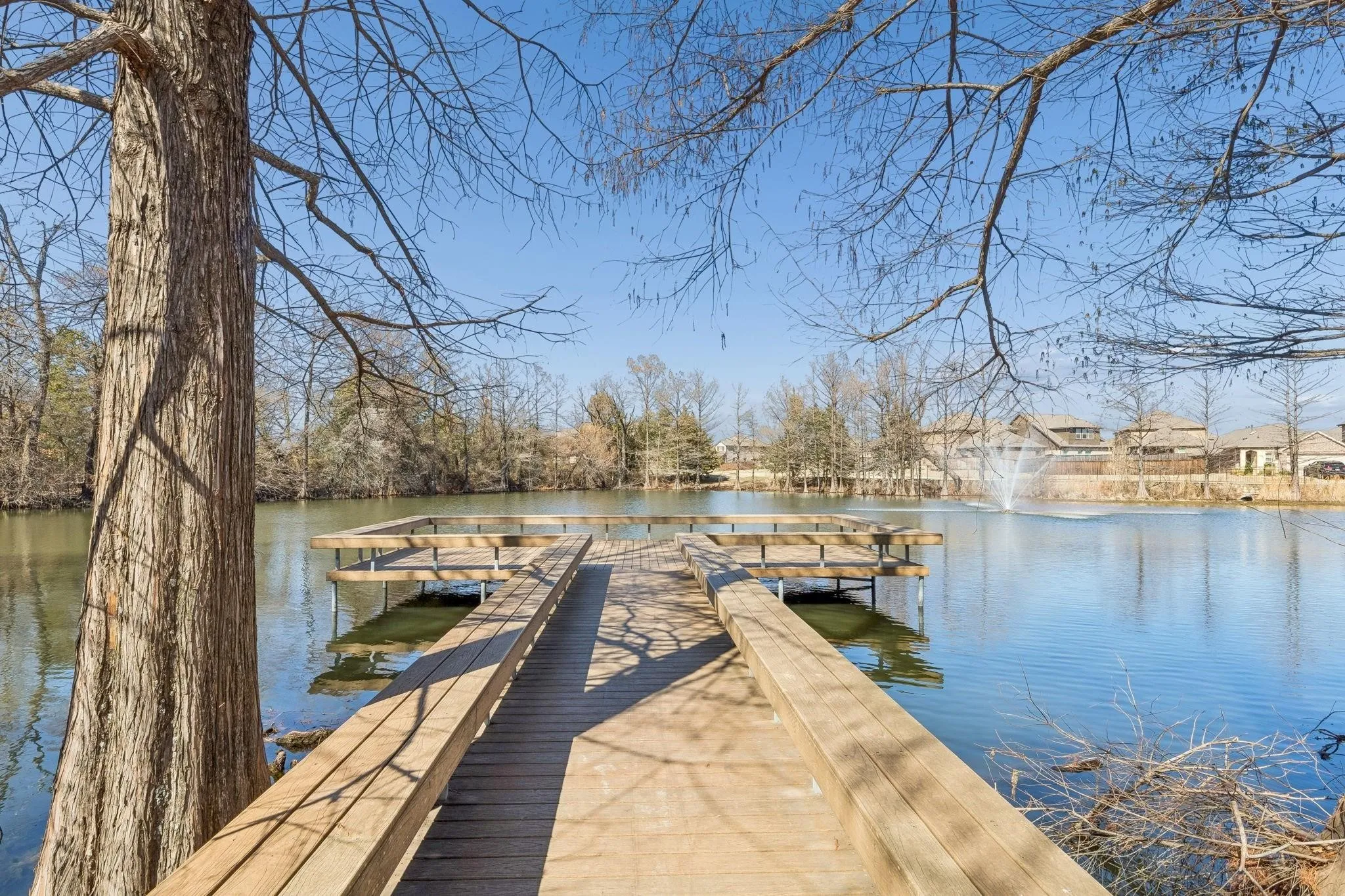 Community dock extending over a placid lake, featuring wood plank construction, built-in seating, and a large fountain in the distance
