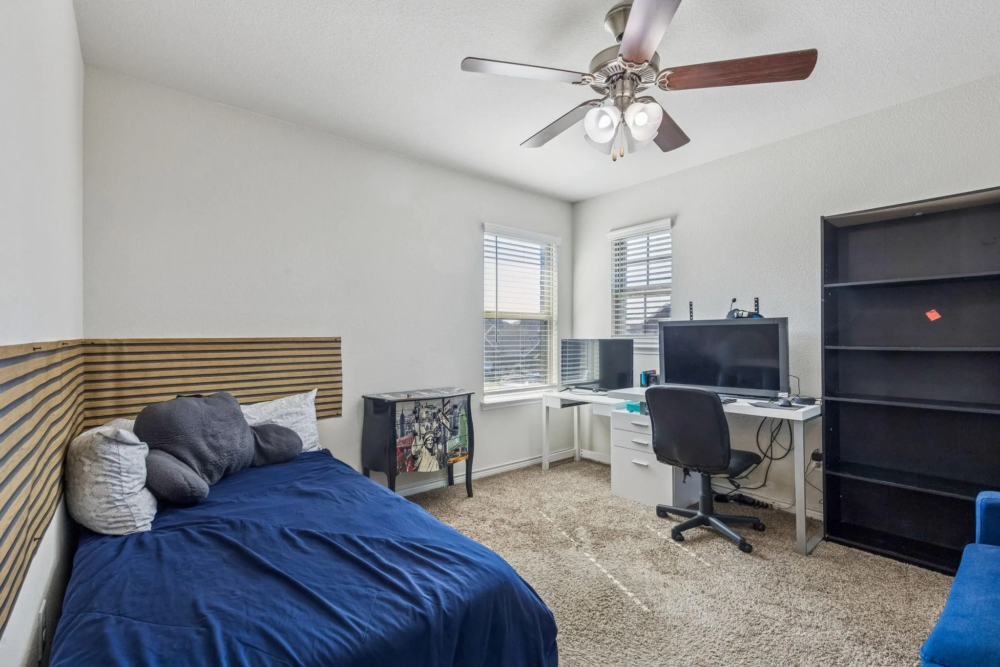 Carpeted room featuring a ceiling fan with light kit, dual windows with blinds, a built-in wooden slat wall detail, and a built-in shelving unit