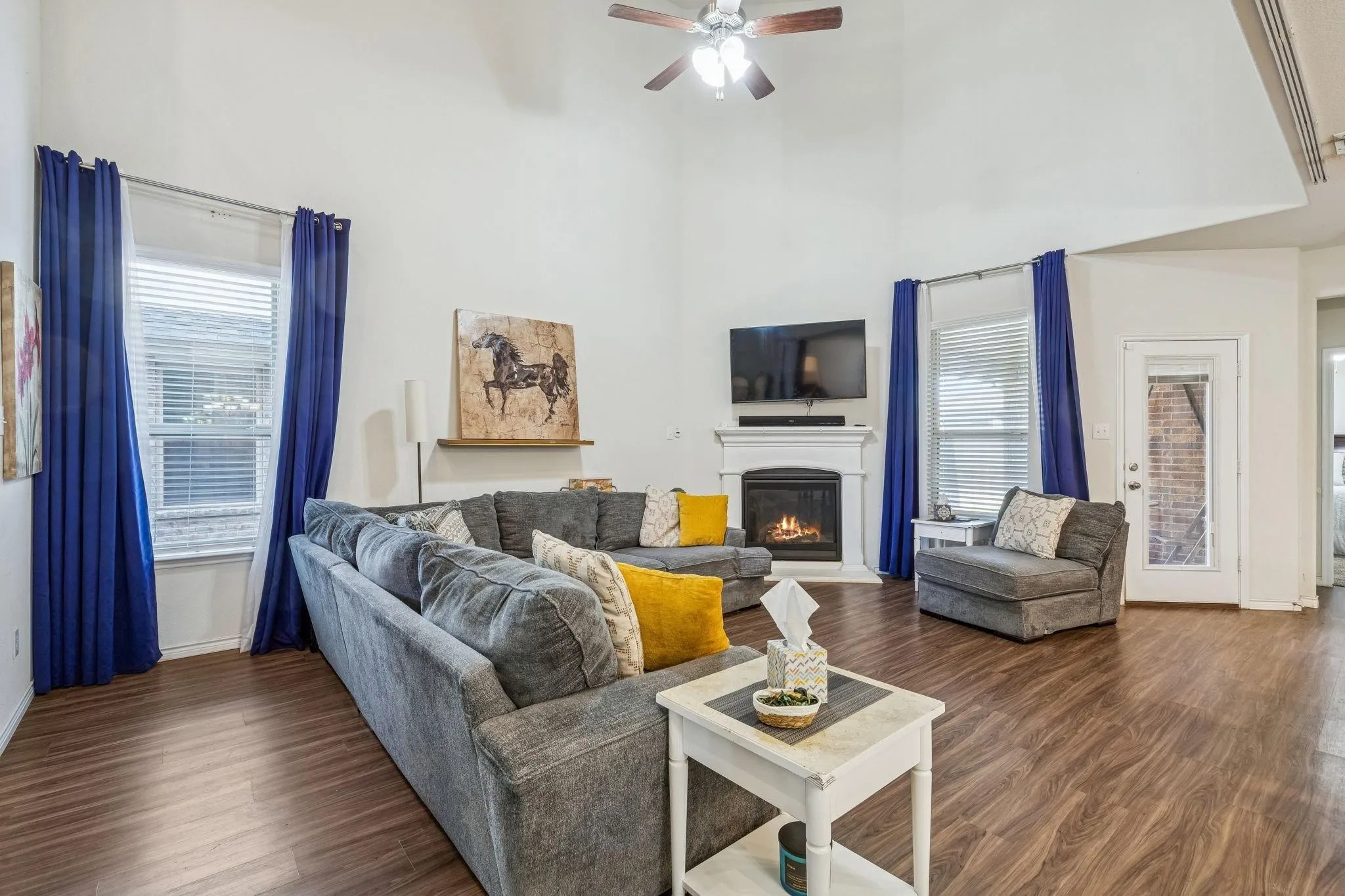 Living area featuring vaulted ceilings, wood-finish flooring, a white mantel fireplace, multiple windows with blinds, and a ceiling fan with light fixture