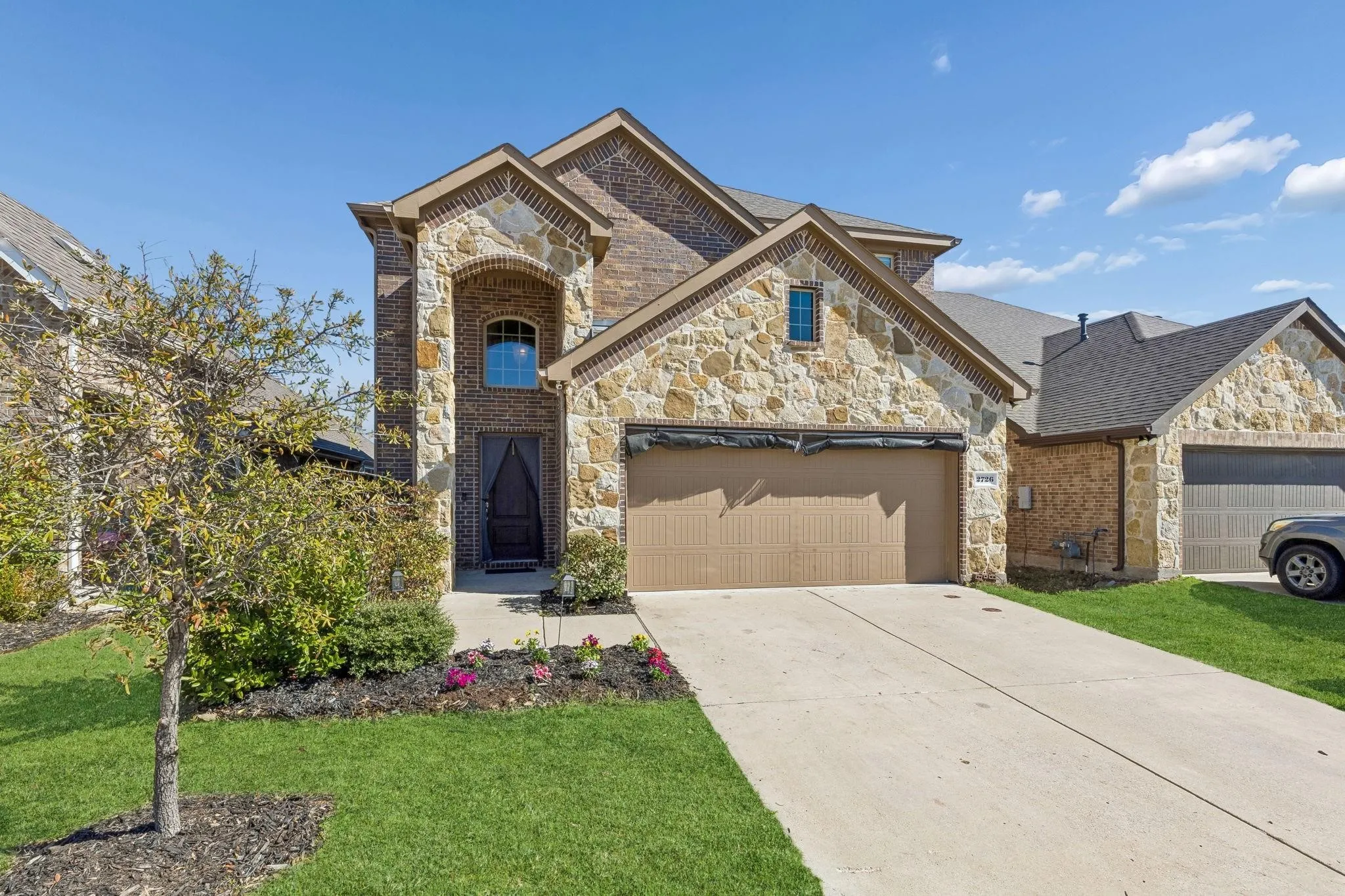 Two-story residence featuring a stone and brick facade, an arched entryway with a dark wood door, a two-car garage with a brown door, a concrete driveway, and landscaped front yard