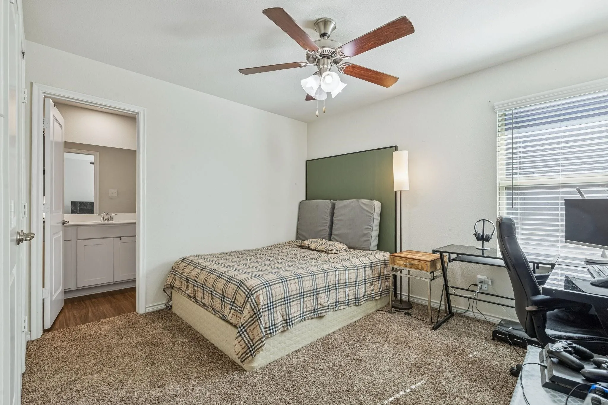 Carpeted room featuring a ceiling fan, window with blinds, and a doorway opening to a bathroom with an ensuite vanity and mirror