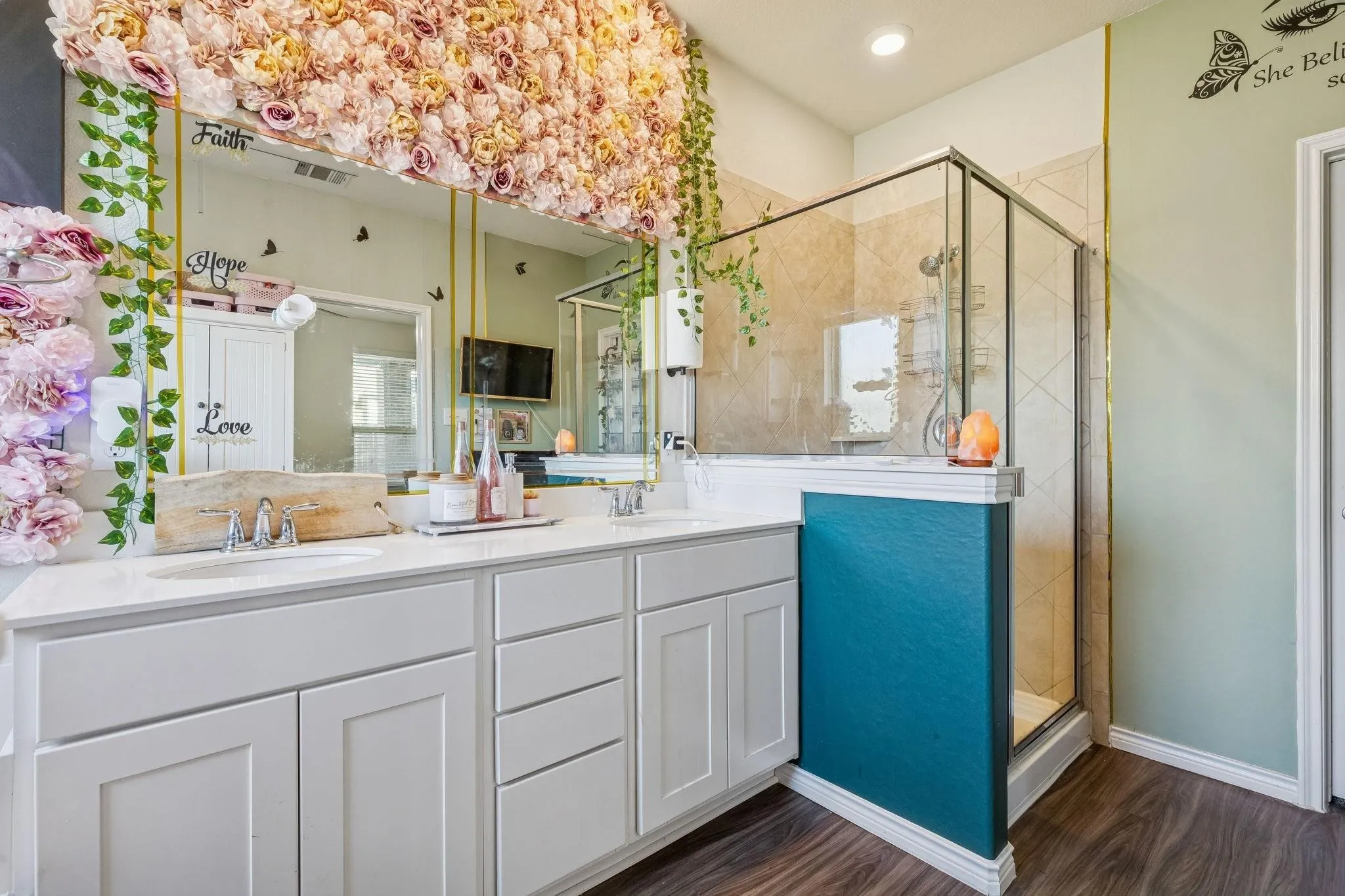 Dual sink vanity with white cabinetry and a light-toned countertop, complemented by a glass-enclosed shower with tiled walls and dark wood-finish flooring