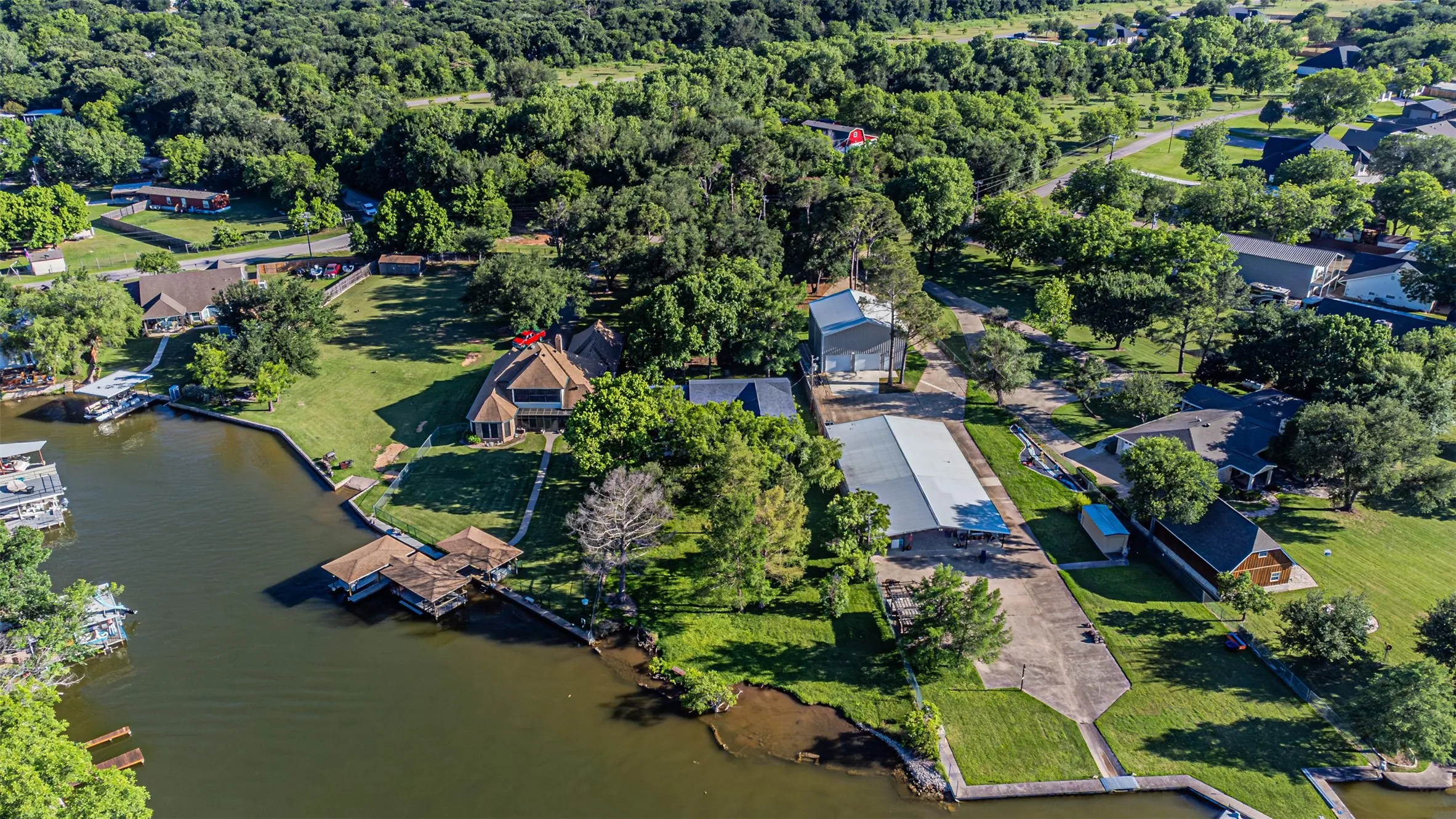 Waterfront property featuring multiple boat docks, a large main residence with an angular roofline, an expansive grassy yard, and mature trees throughout