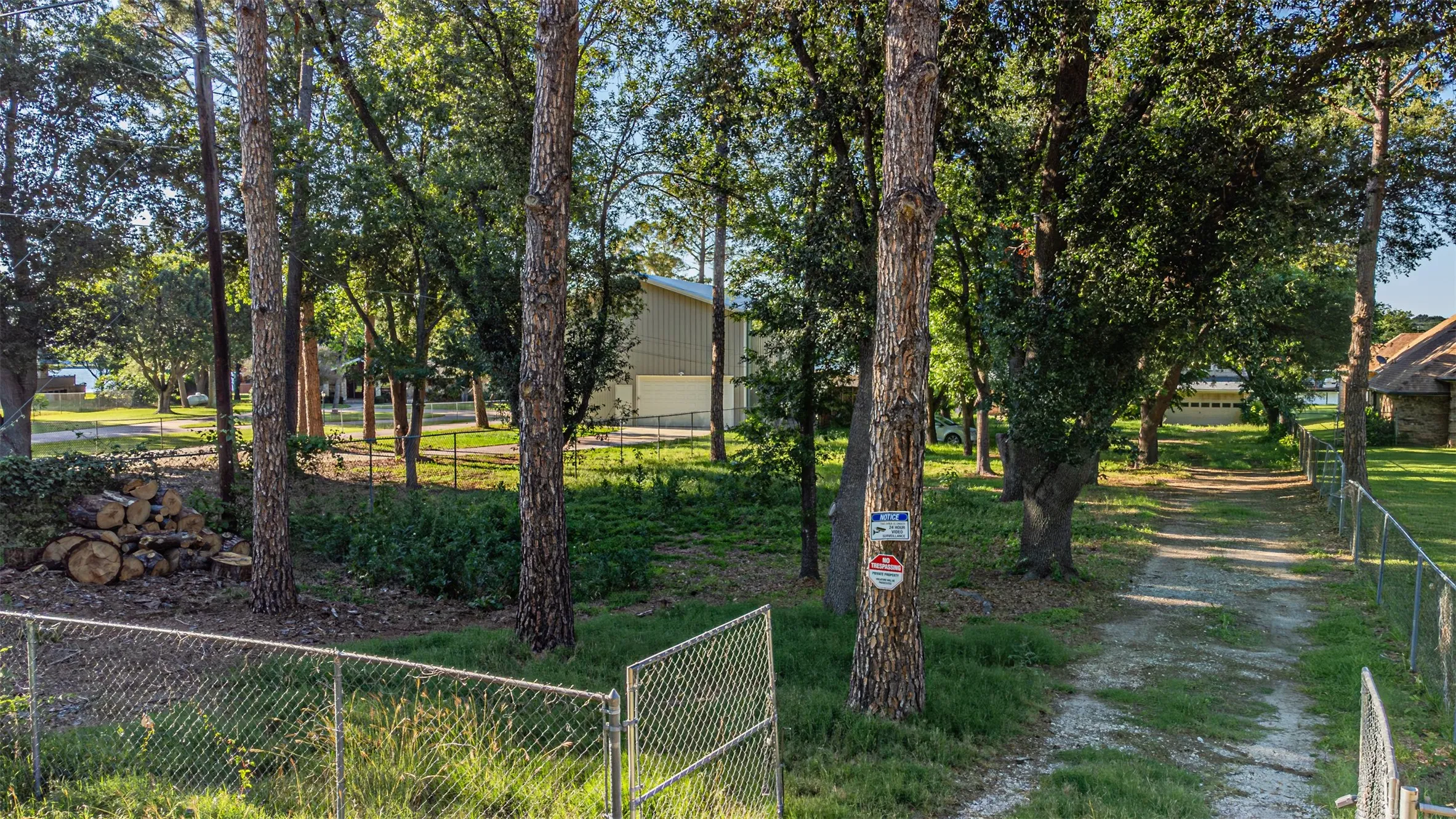 Gated property entrance with a chain-link fence, mature trees, a gravel drive, and natural landscaping