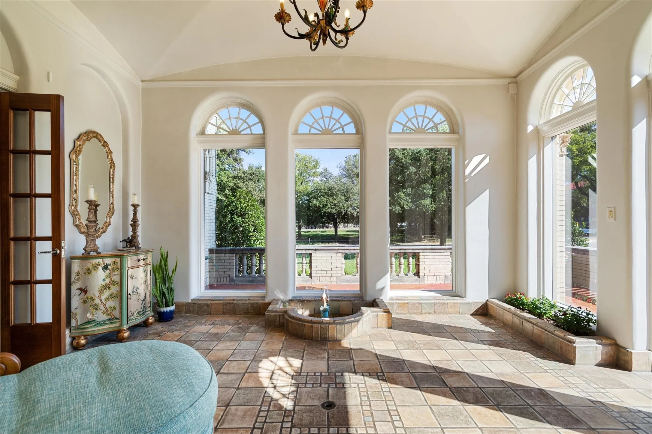 Sunlight pours into the sunroom, where a vaulted ceiling and sparkling chandelier rise above stone
tile floors, while a tranquil water fountain adds movement and charm to the space.