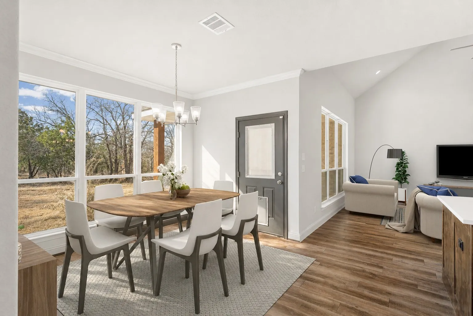 The dining area showcases large windows that bring in abundant natural light and offer serene outdoor views. A contemporary chandelier centers the space, while crown molding and warm wood-look flooring add a touch of elegance.