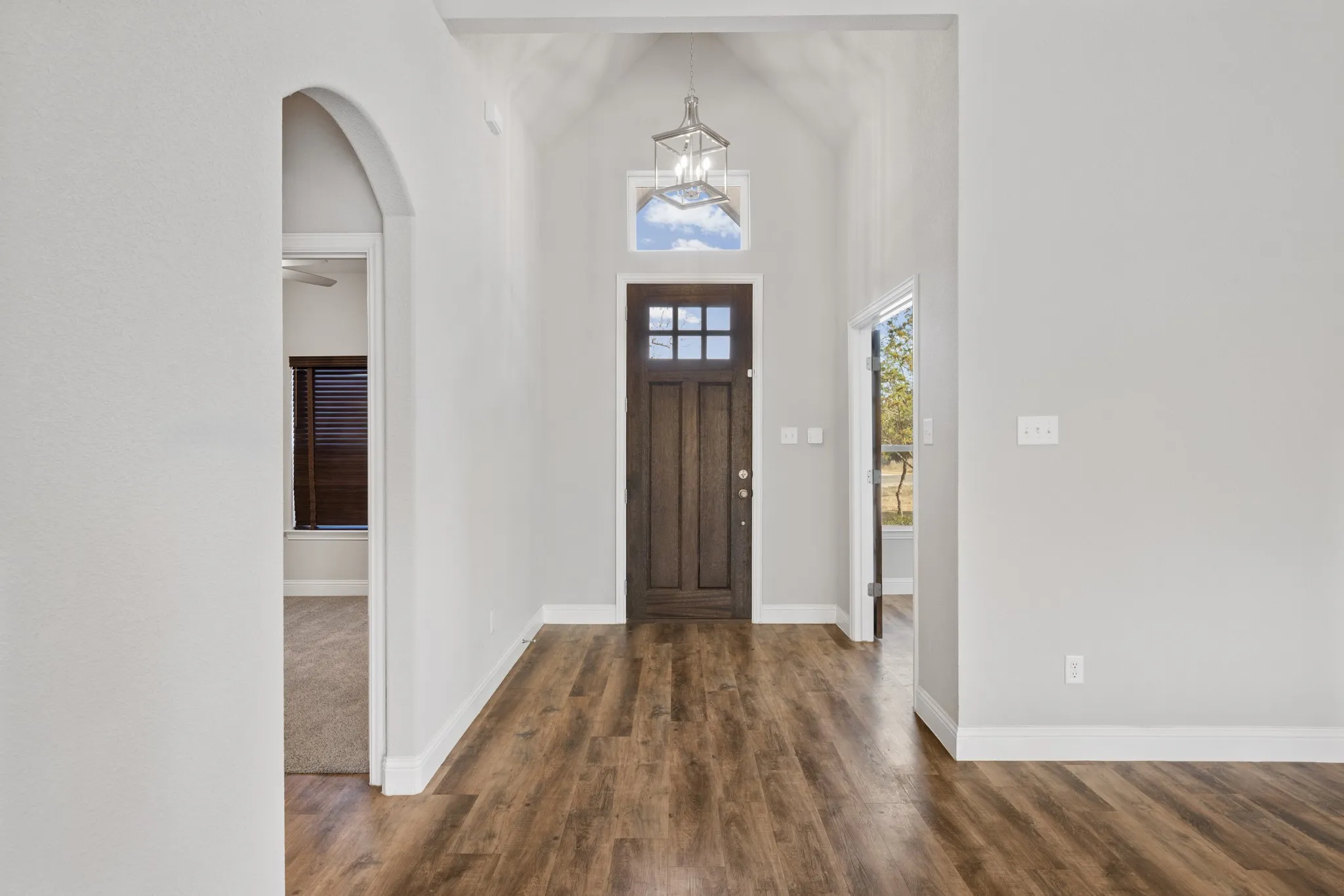 This bright and inviting entryway features soaring vaulted ceilings and a modern lantern-style chandelier that creates an airy, open feel. Warm wood-look flooring flows throughout the space, complementing the rich-toned front door with transom window that fills the foyer with natural light.