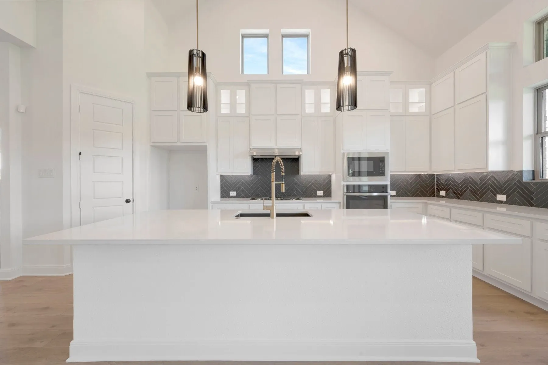 Contemporary kitchen featuring a large central island with an undermount sink, white cabinetry, integrated stainless steel appliances, and a dark herringbone tile backsplash