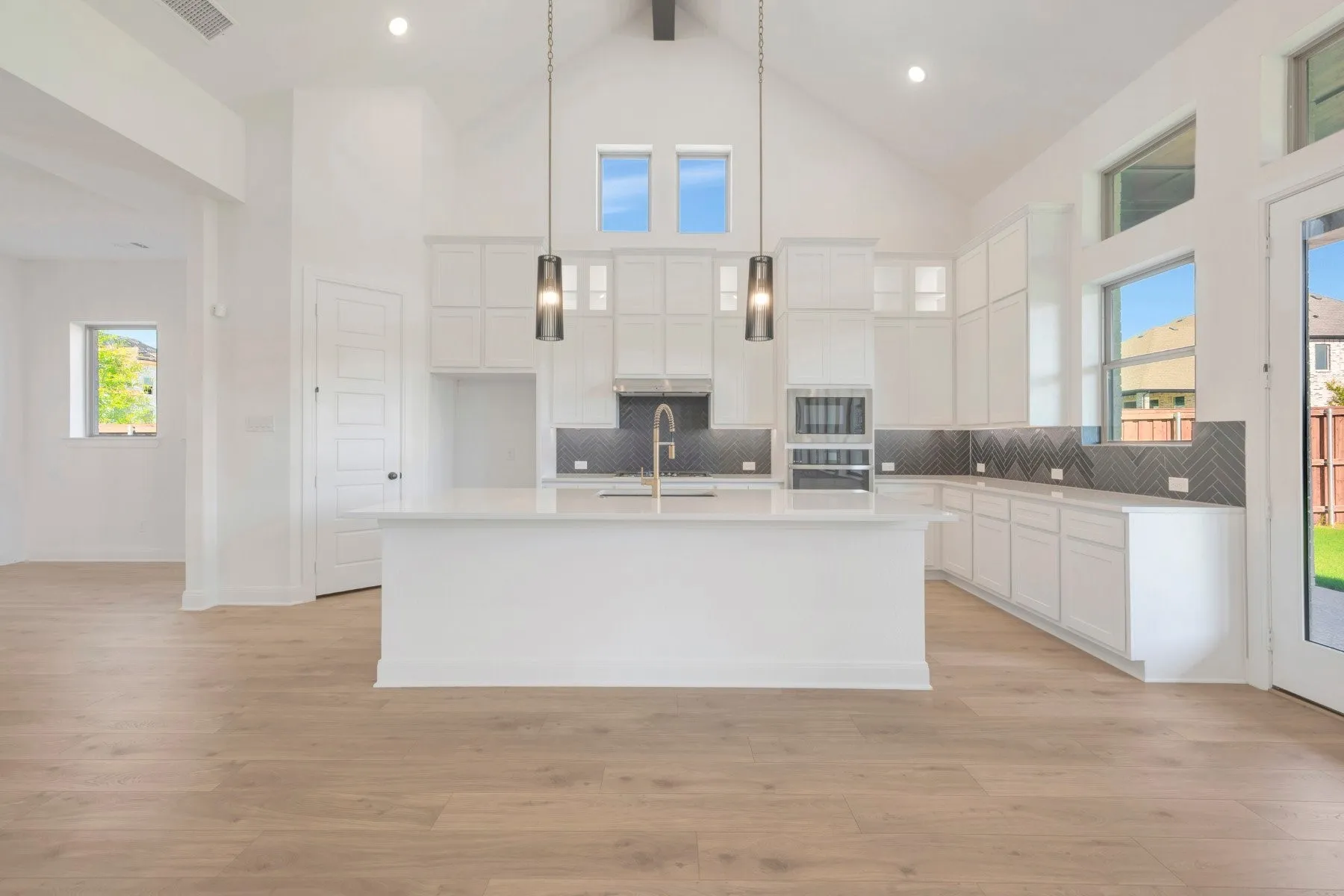 Modern kitchen featuring a large central island, light wood-finish flooring, white cabinetry, dark herringbone-patterned backsplash, and stainless steel built-in appliances