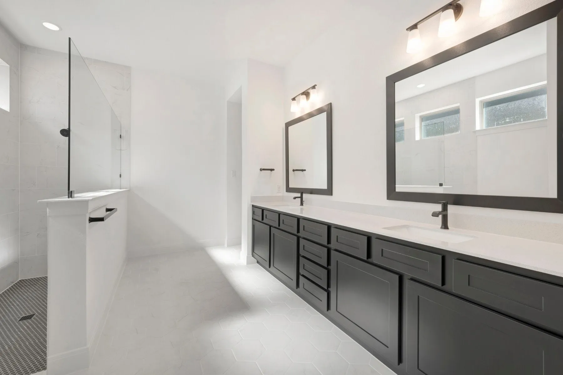 Contemporary bathroom featuring a spacious walk-in shower with a glass panel and dark-tiled floor, dual vanities with dark cabinetry and light countertops, two framed mirrors, and modern light fixtures